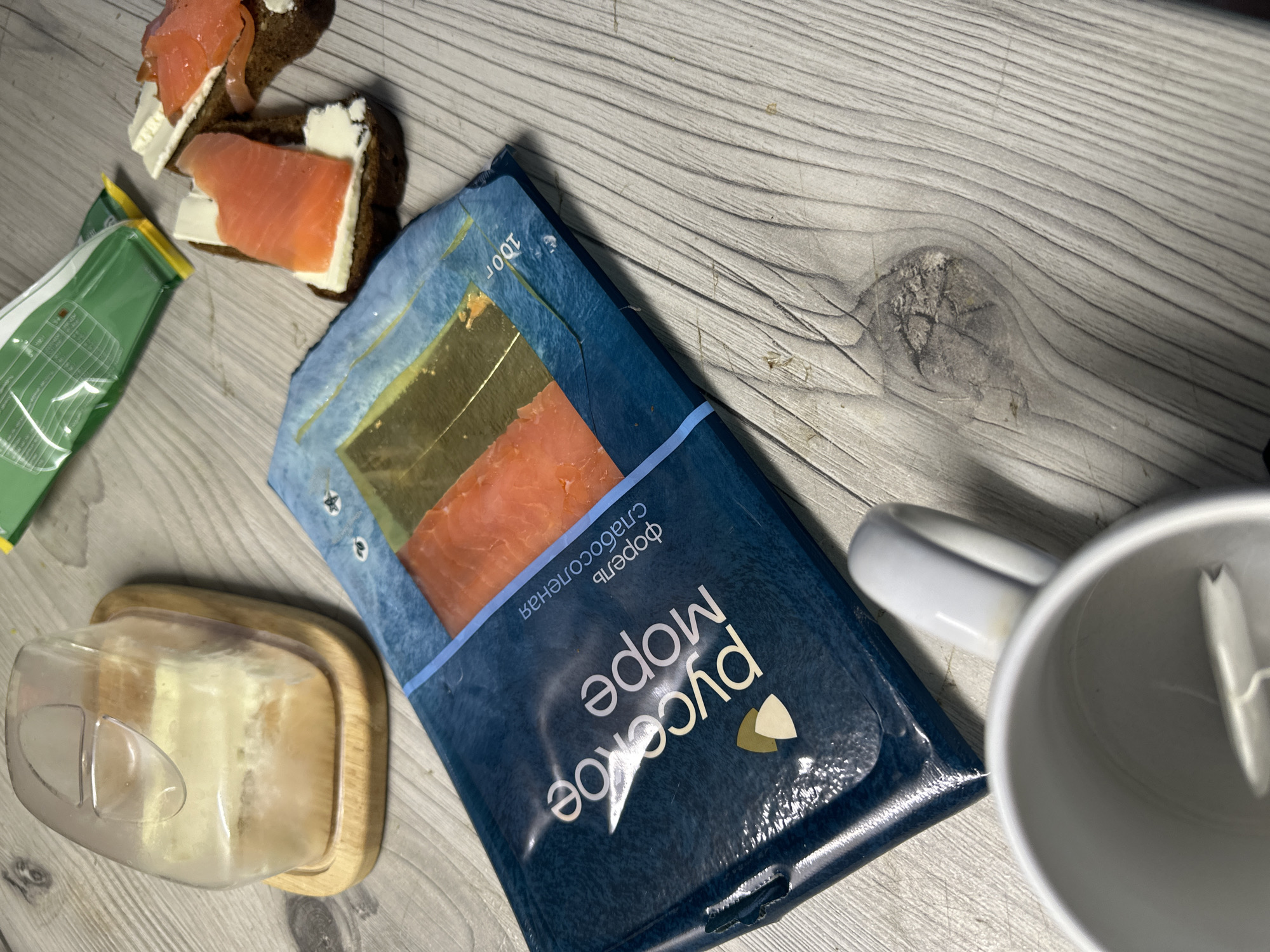 The image shows a still life scene on a light gray wood-patterned surface. A package of "Русское море" (Russian Sea) sliced salmon is prominently displayed, partially open, revealing the fish. Next to it are two open-faced sandwiches made with dark bread, cream cheese, and salmon. A butter dish with butter is also visible. A white mug, possibly containing a spoon, sits to the right of the salmon package. A green package is visible in the periphery, its contents unclear.
The location, Chaikovsky, Russia, is inferred from the Russian language packaging of the salmon.