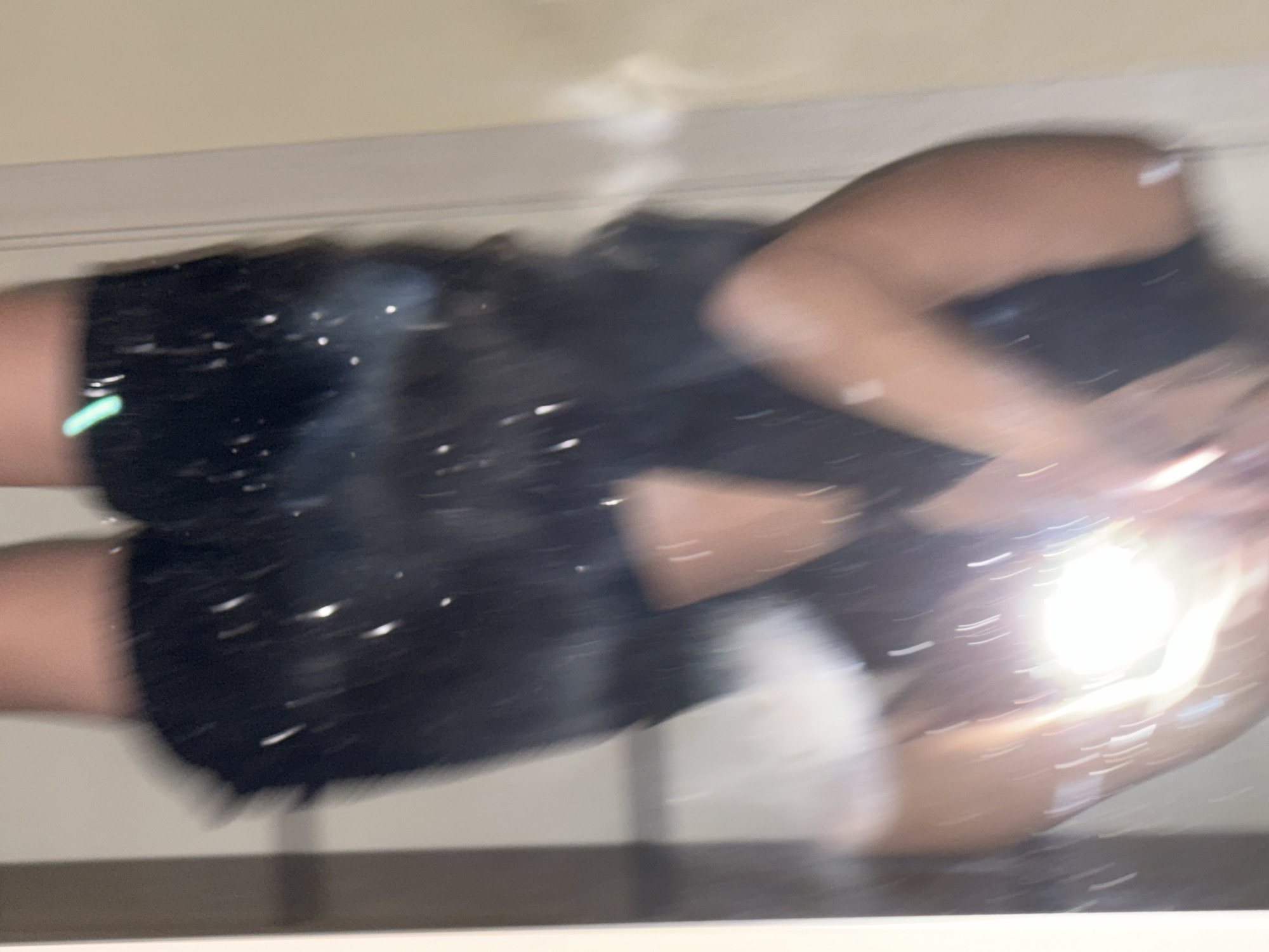 The image shows a person in black clothing, possibly shorts and a top, reflecting in a mirrored surface. The person appears to be holding a phone or camera, indicated by the bright flash. The setting is indoors, suggested by the light-colored wall in the background. There is no information in the image to suggest a specific location, let alone the city of Izhevsk, Russia.
