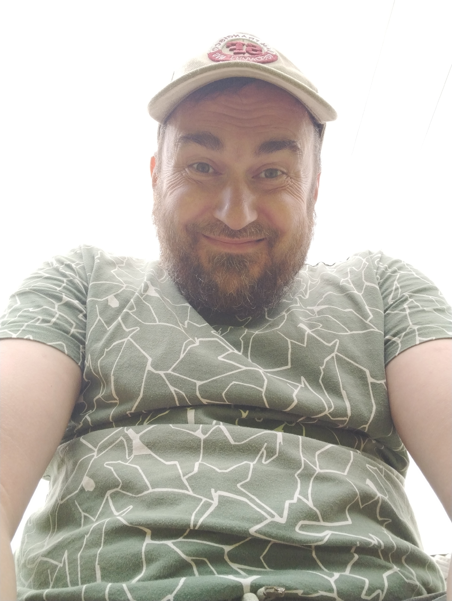 The media file is a close-up, low-angle selfie of a man outdoors, against a bright, featureless sky. He is wearing a light-colored baseball cap with a circular emblem on the front and a green t-shirt with a white, irregular geometric pattern. The man has fair skin, a beard, and appears to be smiling with his eyes slightly closed. The lighting suggests it is daytime, and the bright sky could indicate sunny weather. There are no discernible location cues other than the provided context of "Shiryayevo (Il'inskiy), Russia." The man's posture and the low camera angle imply he is looking up. The overall impression is casual and relaxed, with the man engaging directly with the camera.