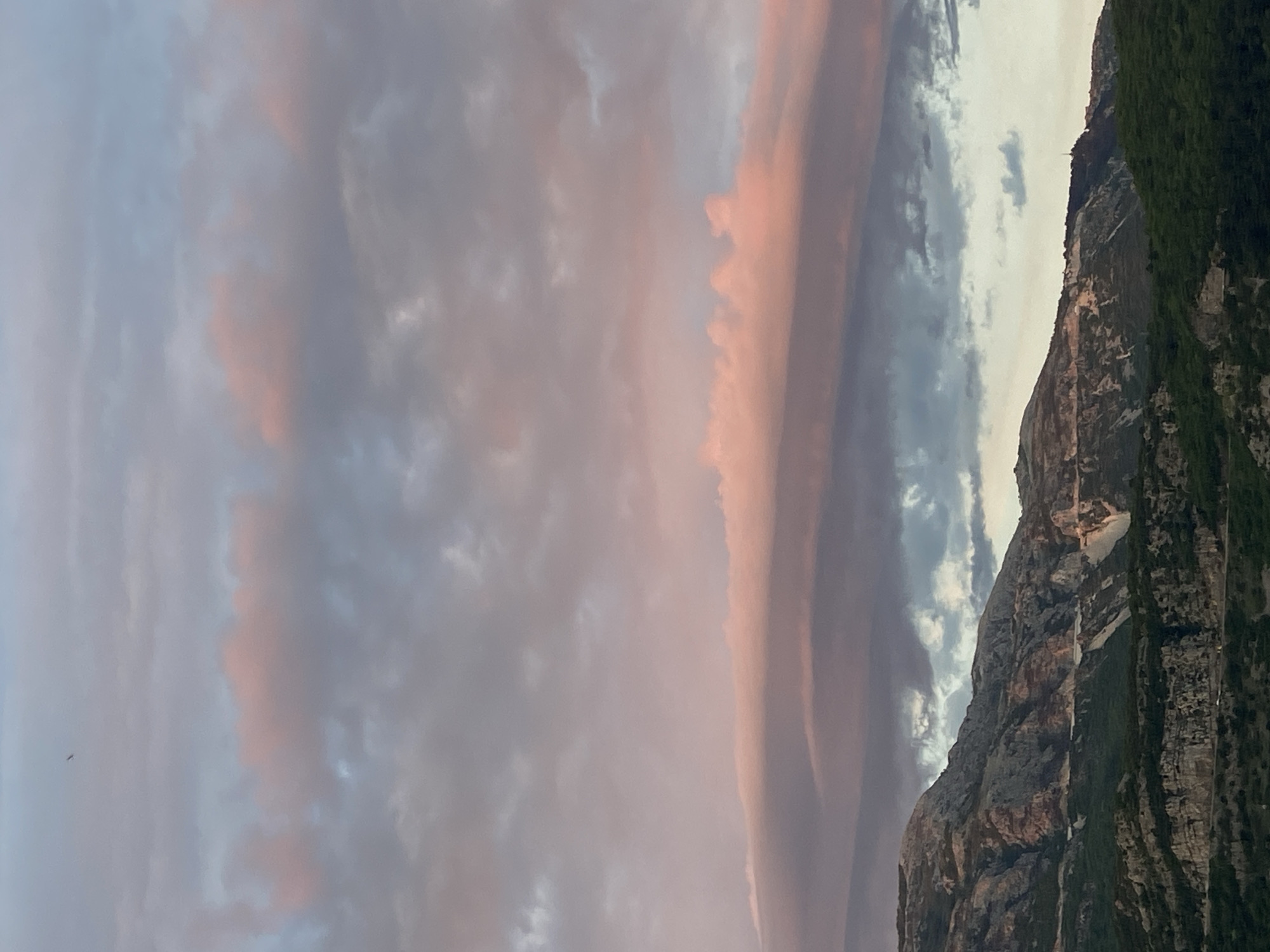 The image presents a majestic mountainous landscape under a dramatic and colorful sky, likely at sunrise or sunset. The sky, filling the majority of the frame, showcases a vibrant mix of soft blue-grey clouds and striking pink and orange hues, particularly pronounced on the right side, indicating the warm glow of low-angle sunlight. The clouds vary from diffuse and wispy formations to more distinct, layered bands.
A rugged mountain range dominates the right side of the composition, appearing as a dark and imposing silhouette against the illuminated sky. The mountains feature steep, rocky terrain with exposed rock faces and noticeable patches of dense green vegetation, especially on the lower slopes and along the ridge lines. A tiny, indistinct structure, possibly an antenna or a marker, is visible on one of the higher peaks. A small, dark speck, perhaps a bird or a distant object, can be seen in the far left of the sky.
The scene evokes a sense of natural grandeur and tranquility, capturing the interplay of light and shadow across the mountainous terrain and the dynamic cloudscape. Given the location context of Budva, Montenegro, these are likely the impressive coastal mountains of the Dinaric Alps. There are no people, explicit activities, or visible text in the image.