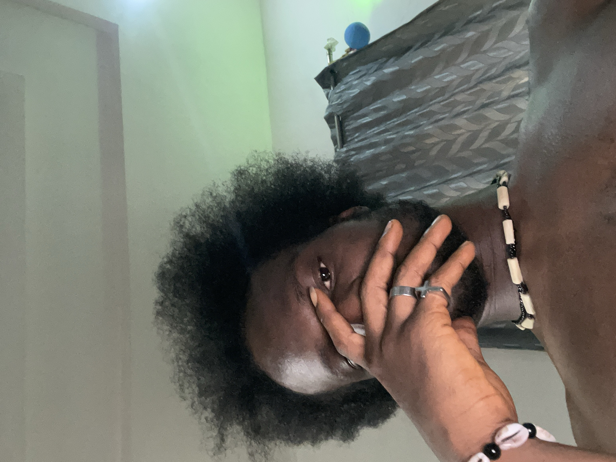 A close-up portrait shows a Black man with an afro covering part of his face with his hand. He has two rings on his fingers. He is wearing a beaded necklace and a seashell bracelet. The background includes a wall, a window with partially closed blinds, and a blue object on the windowsill. The location is in Kuchingoro, Nigeria.