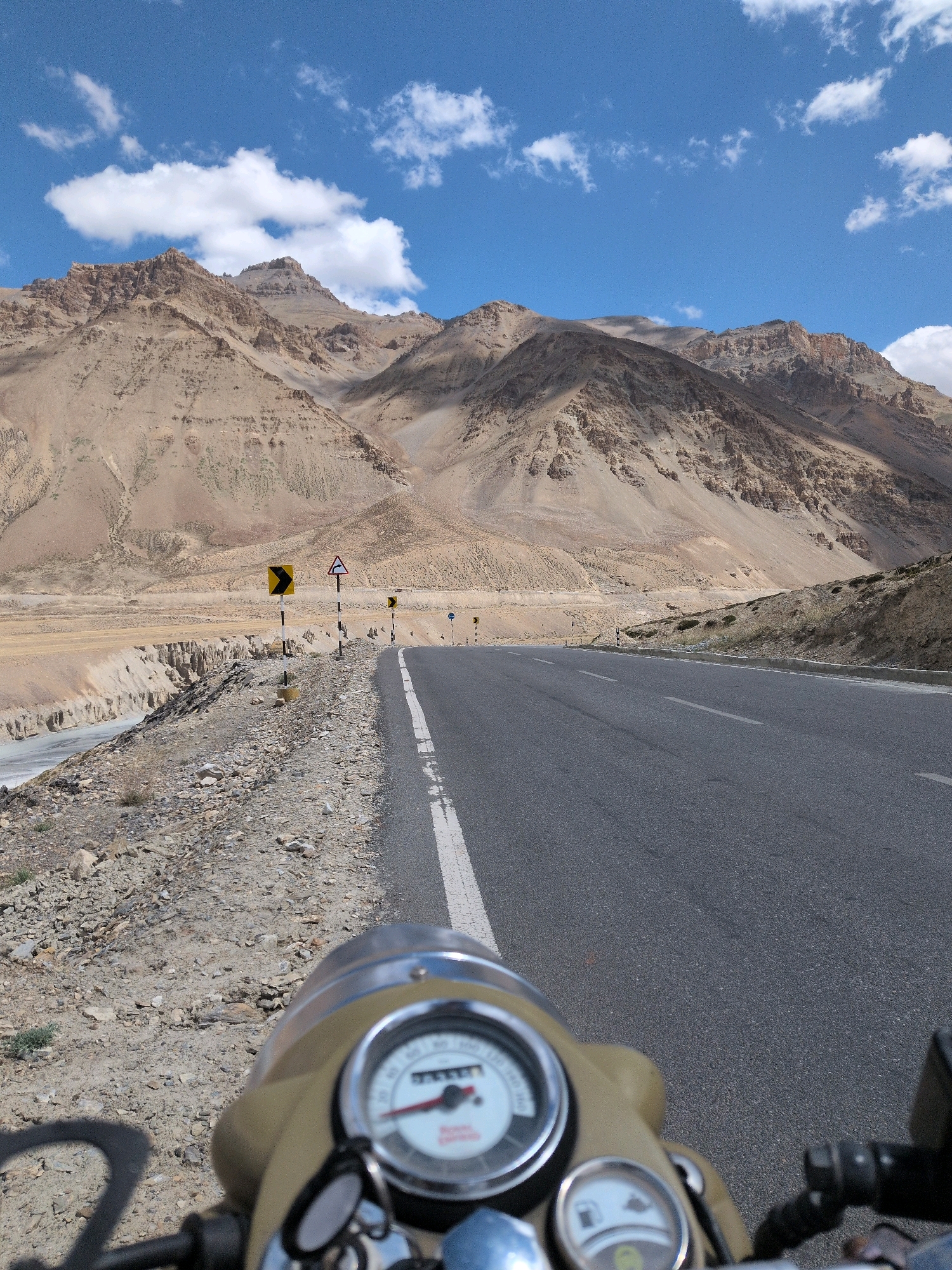 An eye-level, first-person perspective shot shows a motorcycle dashboard and handlebars in the foreground, with a winding asphalt road stretching into the distance. The road is bordered by a rocky, arid landscape on the left and a more gently sloping, vegetated hillside on the right. Majestic, barren mountains with jagged peaks dominate the background under a bright blue sky dotted with white clouds. Road signs are visible along the left side of the road. The scene appears to be in a remote, mountainous region, possibly a highway in Ladakh, India, given the characteristic landscape and road markings. There are no people visible in the image.