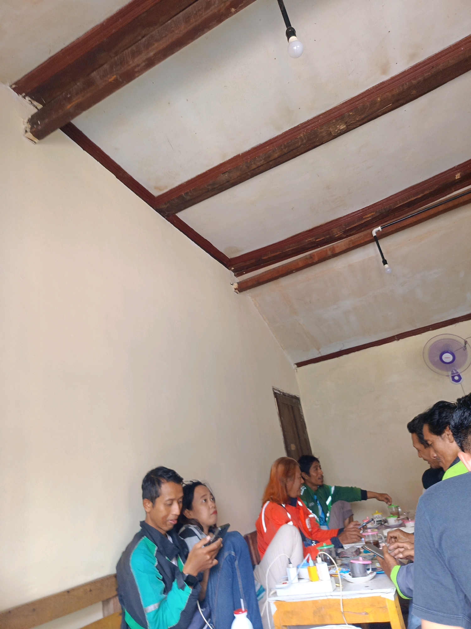The image depicts an indoor scene in Kebonagung, Indonesia, likely a humble gathering place, possibly a home or a small communal area. The room has a rustic aesthetic with exposed wooden beams forming the ceiling structure. The walls are a neutral, light color, and the lighting appears to be a mix of ambient light and a bare bulb hanging from the ceiling.
In the foreground, a man in a teal and black jacket is seated, looking at his phone. Next to him, a woman in a denim jacket is also looking at a phone. Further back, a woman with reddish-brown hair, wearing an orange jacket, is seated and looking towards the right side of the frame. To her right, a man in a green jacket is seated and appears to be interacting with objects on a table. Several other individuals are visible in the background, suggesting a social gathering.
On a table in the mid-ground, there are various items including cups, a kettle, and electrical equipment, indicating that food and drinks are being served or prepared. A small fan is mounted on the wall in the upper right corner of the image. The overall impression is one of a casual, informal gathering of people.