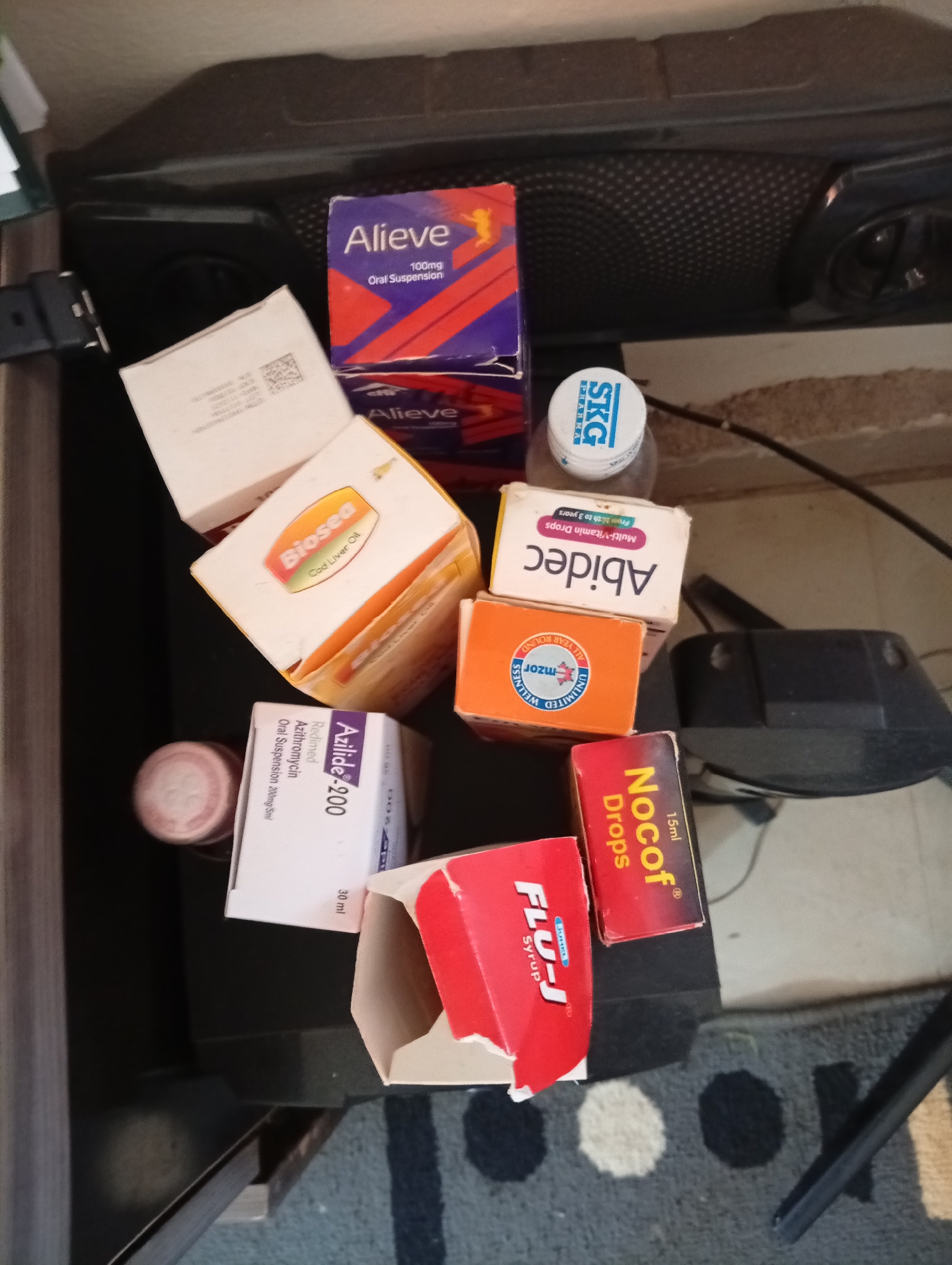The image is a close-up overhead shot of various medicine boxes and bottles arranged on a dark surface, possibly a table or shelf. The objects are clustered together in the center of the frame. The medicine boxes include "Alieve," "Biosea Cod Liver Oil," "Abidec Multi-Vitamin Drops," "Azilide-200 Oral Suspension," "Nocof Drops," and "Flu-J Syrup." A bottle with an "SKG Pharma" cap is also visible. The background is dark, with some texture suggesting a speaker grille. The lighting is artificial and somewhat harsh, casting shadows on the objects. There is no indication of the specific location within the city of Gura Top, Nigeria, beyond it being an indoor setting. No people are present in the image.