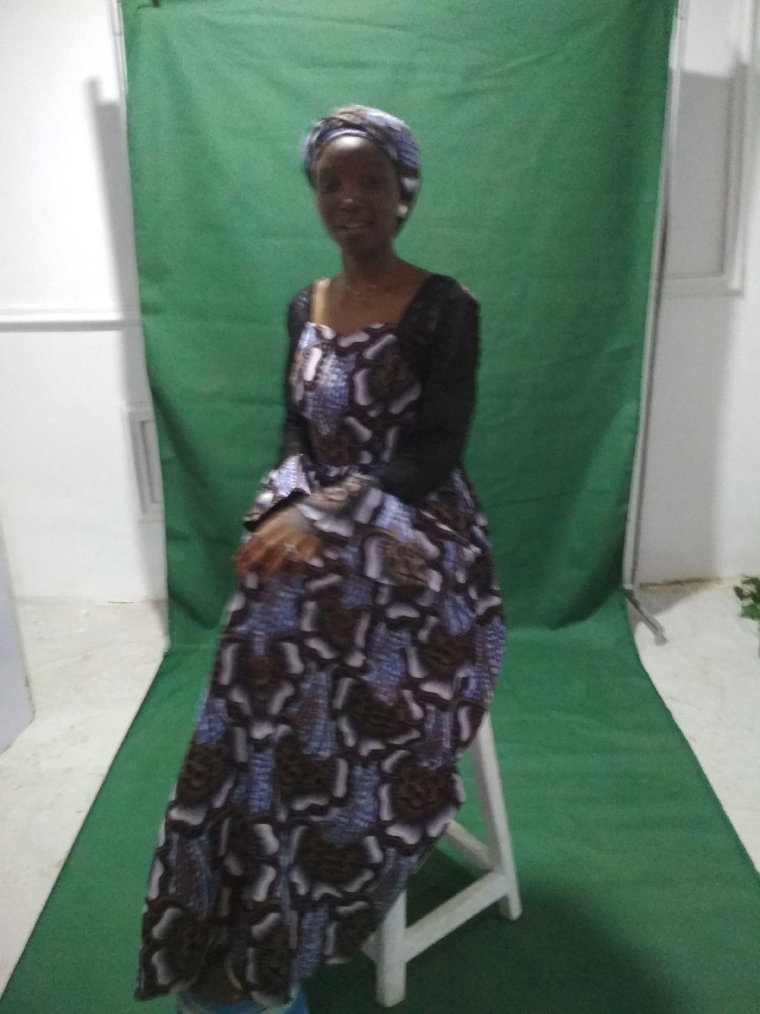 A woman sits on a white stool in front of a green backdrop. She is wearing a long dress with a head wrap. The background consists of a green cloth draped as a backdrop and flooring. A white wall is visible on the left side of the image. The location is Unknown, Unknown.