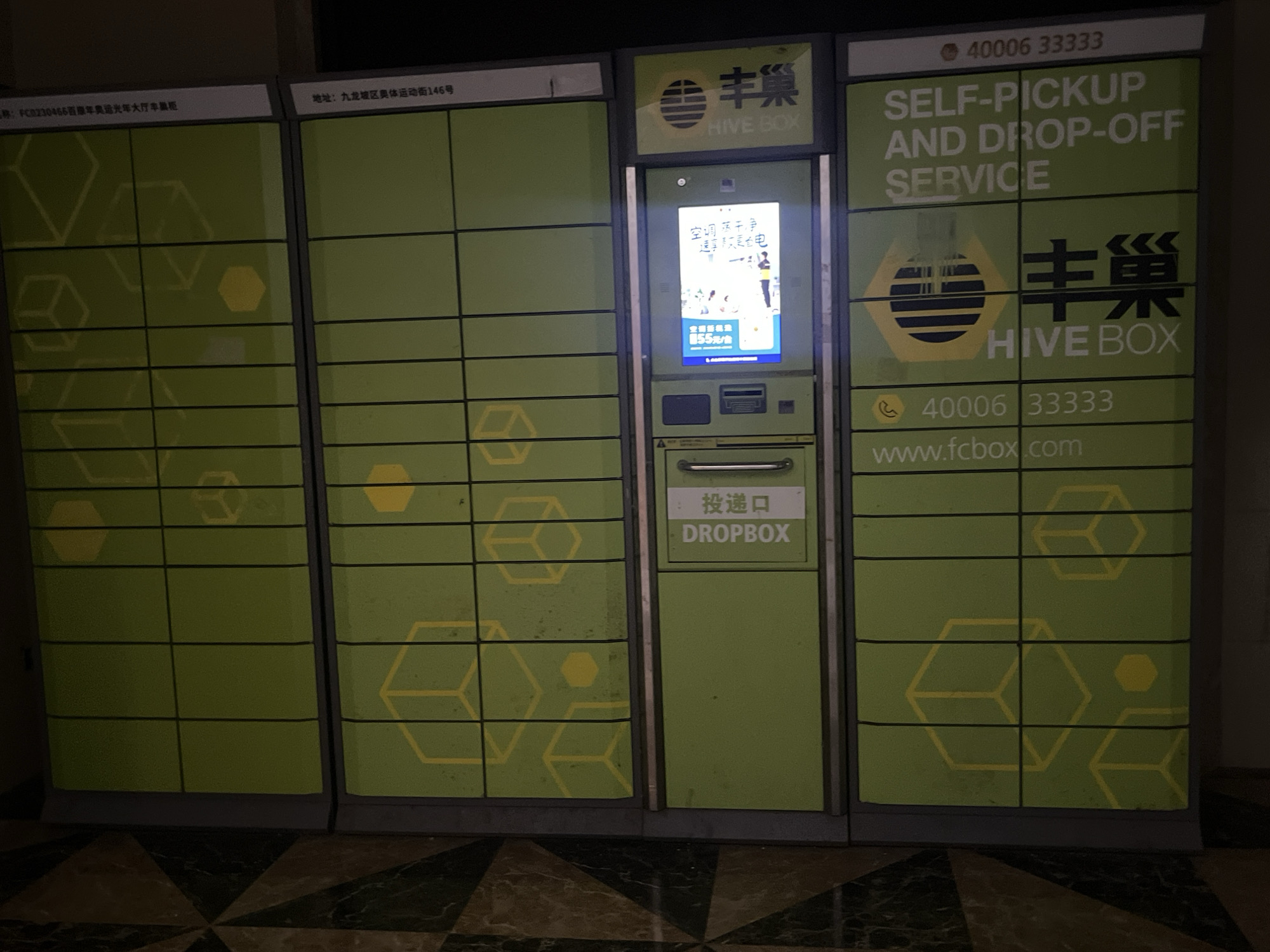 The image shows a row of green self-pickup and drop-off smart lockers, branded "HIVE BOX." The lockers are arranged in a bank, with one unit featuring a digital screen displaying an advertisement and a keypad for user interaction. The advertisement has Chinese text and images of people. Above the lockers, there are signs with Chinese characters and the English text "SELF-PICKUP AND DROP-OFF SERVICE." One sign also displays the phone number "40006 33333" and a website address "www.fcbox.com." A sign on the left side of the lockers indicates a location name in Chinese characters. The lockers are situated against a wall, and the floor in front of them is covered with a patterned tile. The scene appears to be in an indoor public space. The text on the left side of the lockers indicates the location as "Jiulongpo District, China."