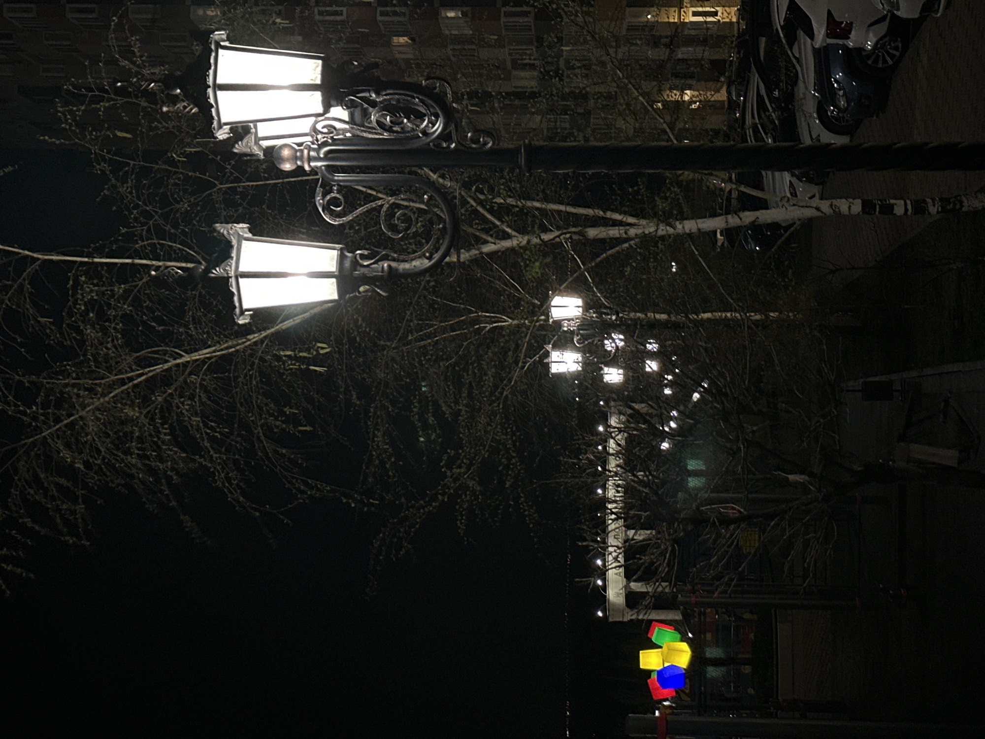 At night in Rostov-on-Don, Russia, a street scene features a vintage-style streetlight with two illuminated lamps. The ornate black metal lamppost is adorned with decorative scrolls. Bare tree branches extend around the streetlight, and further down the street, a line of similar streetlights stretches into the distance. Near the bottom of the frame, brightly colored, illuminated cube-shaped decorations are visible. In the top-right corner, parked vehicles are visible along the edge of a building with illuminated windows.