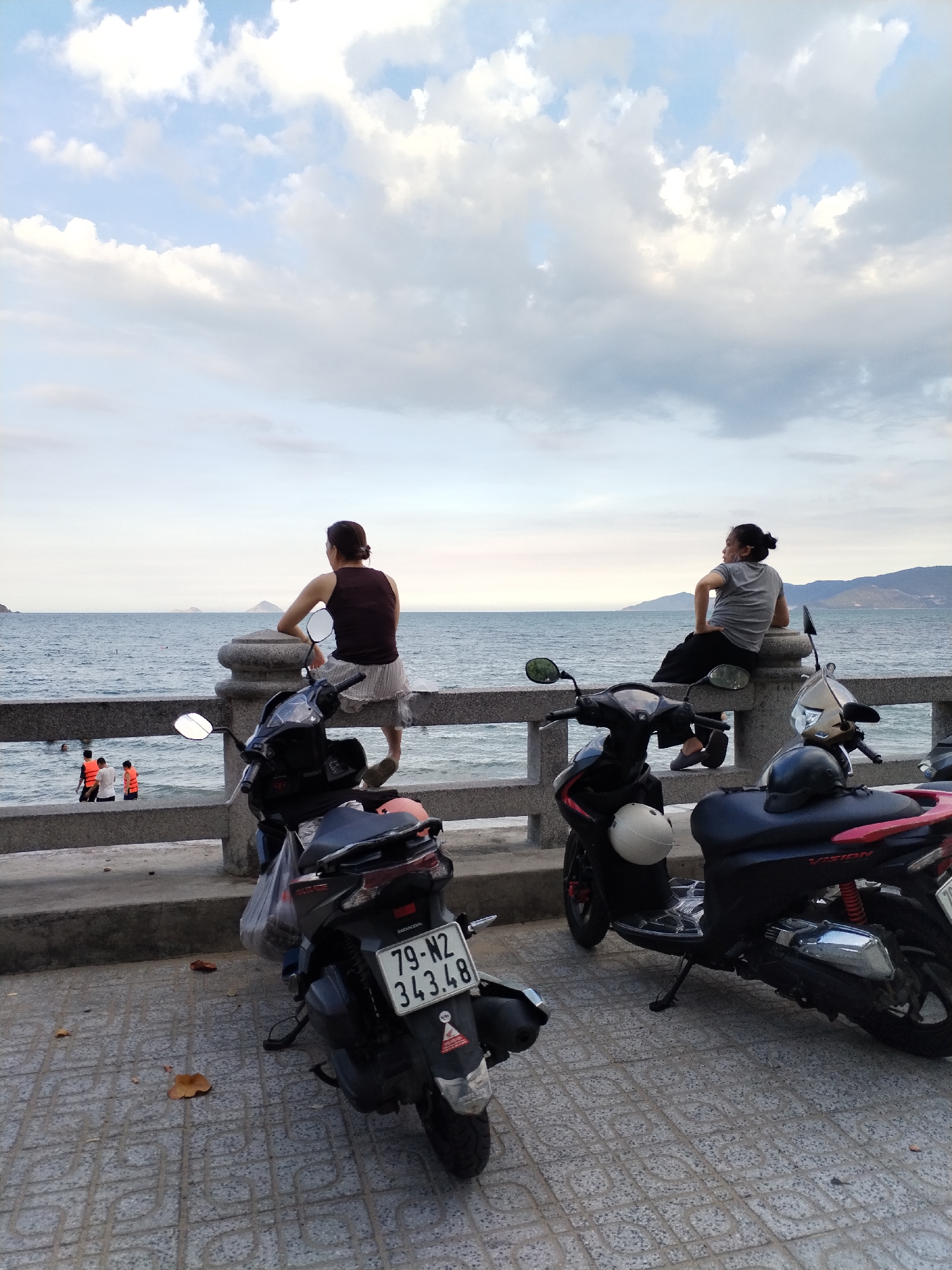 The image depicts a waterfront scene in Bac Nha Trang Ward, Vietnam. Two women are seated on a concrete barrier overlooking the ocean. The woman on the left wears a brown tank top and striped skirt; the woman on the right wears a gray shirt and black pants. Three individuals wearing orange life vests are partially visible in the water to the left. Three parked motorcycles are in the foreground. The motorcycle to the left has a license plate reading "79-N2 343.48". The sky is cloudy. The pavement is tiled.