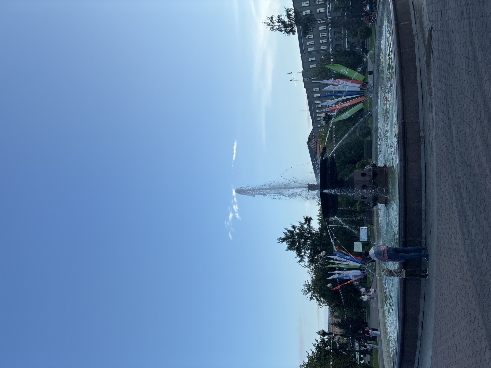 The image depicts a fountain in Kirov Park, Irkutsk, Russia. The fountain is circular with a central spout spraying water upwards, and smaller streams of water arching outwards. Several flags are displayed around the fountain. In the foreground, a person wearing a pink and white striped shirt and blue jeans stands with their back to the camera, looking towards the fountain. A child in a camouflage jacket and green pants stands in front of them, also facing the fountain. In the background, a building with multiple windows is visible, along with trees and lampposts. The sky is clear blue with a few wispy clouds. The ground is paved with brick-like tiles.
