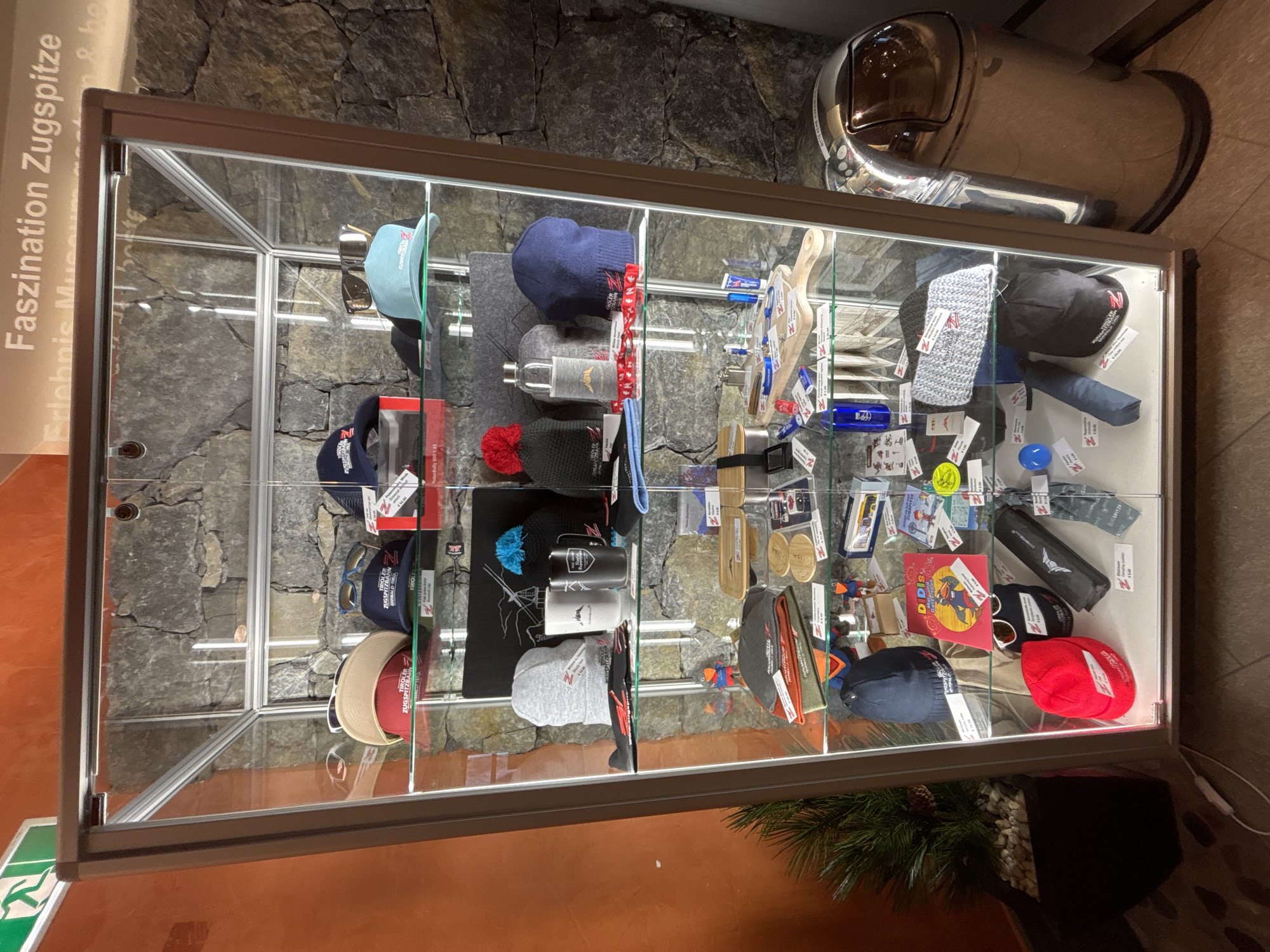 The image displays a glass display cabinet filled with various souvenir items, predominantly hats, sunglasses, and other accessories, likely at the Zugspitze "Erlebnis Museum" in Ehrwald, Austria, as indicated by the text "Faszination Zugspitze Erlebnis Museum" visible at the top of the cabinet. The cabinet is illuminated from within, highlighting the items on multiple shelves. The background consists of a rough stone wall. To the right of the cabinet, a shiny, metallic trash receptacle is visible. The scene is indoors, with a plant in a dark pot to the left of the cabinet. The items in the cabinet appear to be merchandise related to the Zugspitze, an alpine peak.