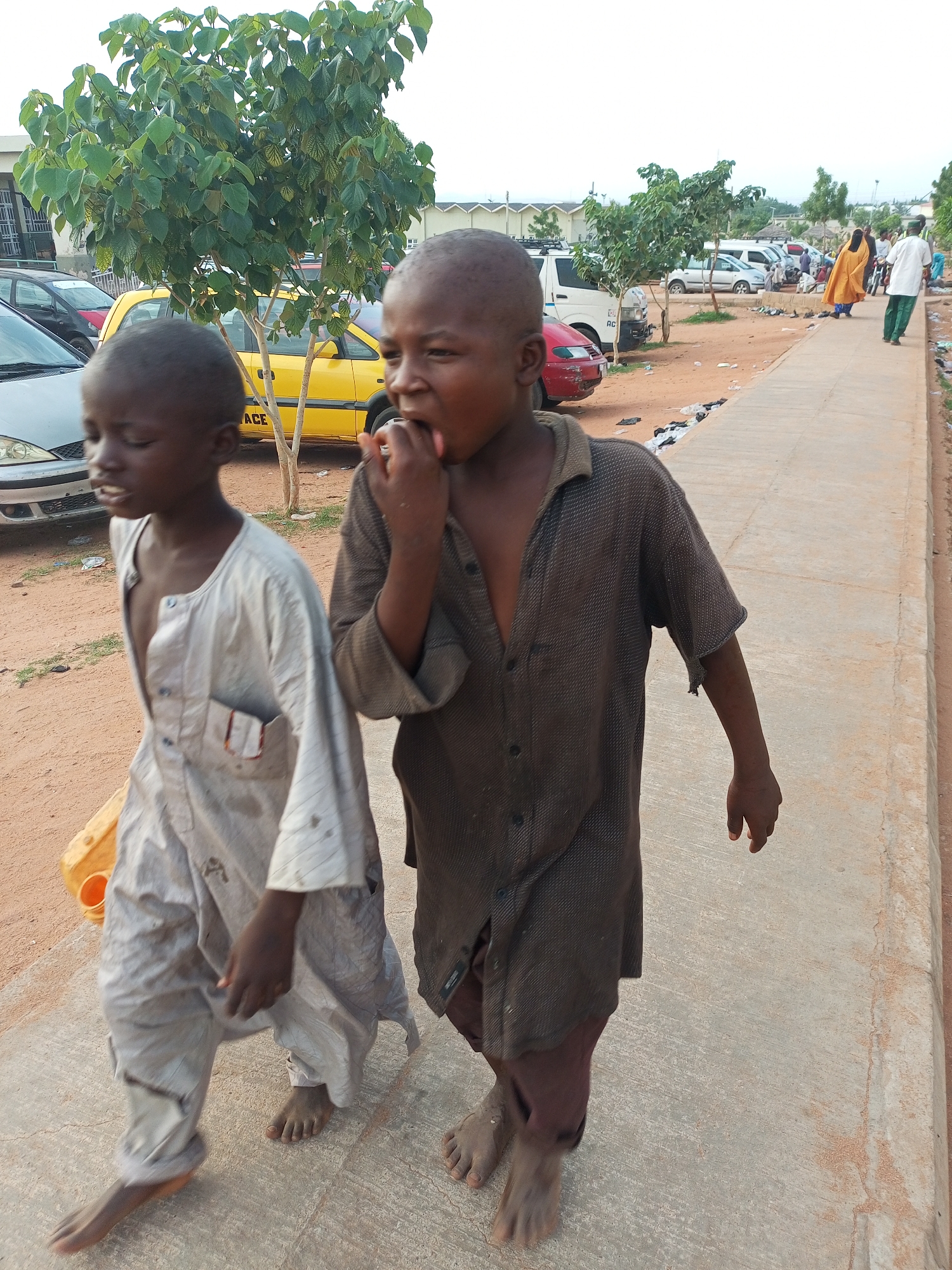 In Gombe, Nigeria, two young boys walk barefoot on a concrete pathway. The boy on the left wears a light-colored, loose-fitting garment and carries a yellow jerrycan. He is looking downwards and his mouth is slightly open. The boy on the right is wearing a dark, button-up shirt that is open at the neck and dark pants. He has his hand up to his mouth, as if eating or touching his lips. Behind them, several parked vehicles, including a yellow bus and white vans, are visible. A lush green tree stands to the left of the frame. The ground around the pathway is sandy and littered with some debris. In the background, other individuals can be seen walking and standing, and buildings are present. The sky is overcast.