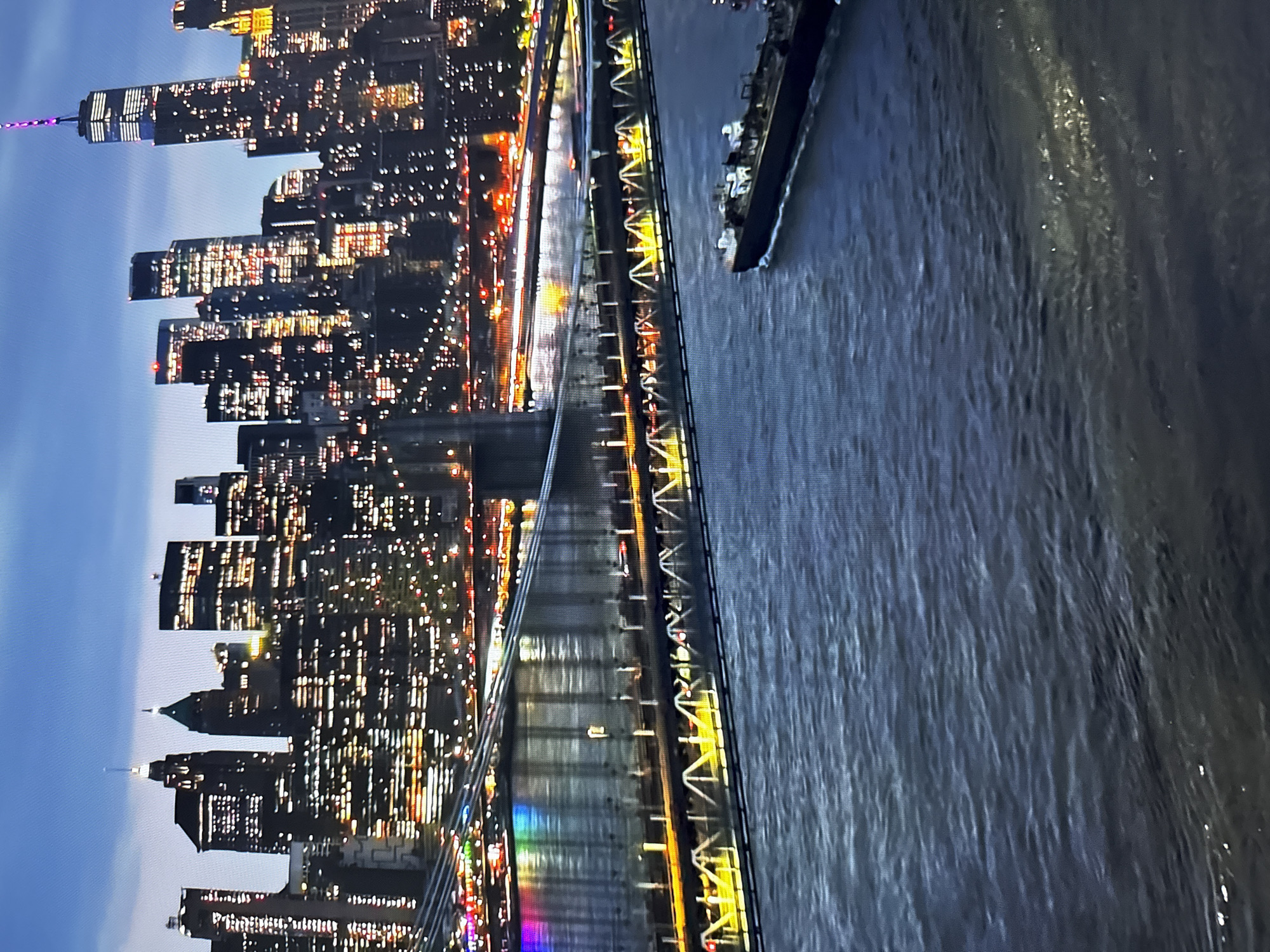 The image displays a wide-angle, high-angle view of a city skyline at dusk, dominated by numerous illuminated skyscrapers. A large bridge, possibly a suspension bridge, stretches across a body of water in the foreground. The bridge has multiple levels and is lit with yellow and white lights. Below the bridge, a dark-colored barge or ship is navigating the water. The water is choppy and reflects the lights from the city and bridge. The sky above is a deep blue, transitioning to darker shades of twilight. The scene is urban and appears to be a major metropolitan area, likely featuring a river or harbor. The city of Burgas, Bulgaria is not depicted in this image; the architecture and scale are more characteristic of a large North American city like New York City.