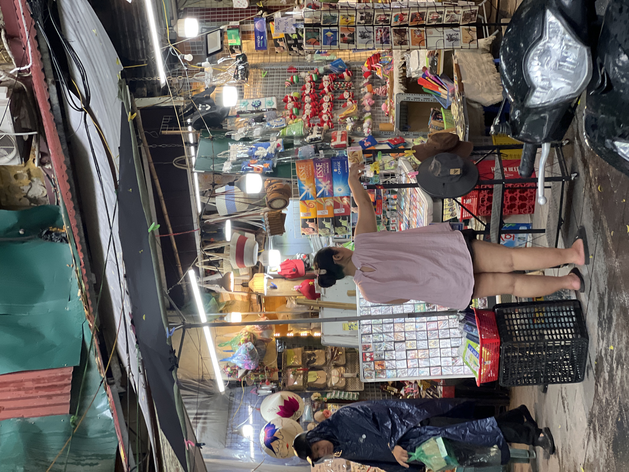 This image depicts a bustling street market scene in Hanoi, Vietnam, likely on a rainy day given the presence of a person in a dark blue hooded raincoat. The market appears to be an outdoor or semi-outdoor stall, with a corrugated metal roof and various awnings providing shelter.
In the foreground, a person wearing a dark blue hooded raincoat and black pants is walking from left to right, carrying a light green plastic bag. To the right of this person, a woman with her back to the camera is wearing a light pink sleeveless top and dark shorts. She is standing with her right arm extended, appearing to interact with a display of goods.
The market stall is filled with a diverse array of products. There are many bags of different types and sizes hanging from the ceiling, including straw bags, canvas bags, and smaller pouches. In the center, a rack displays numerous small, festive Christmas-themed ornaments, possibly Santa figures. There are also displays of what appear to be postcards or small prints, as well as various other trinkets and souvenirs. A red basket filled with colorful items, possibly pens or small boxes, sits atop a black plastic crate to the left of the woman in the pink top.
A black motorcycle is parked in the lower right portion of the image, partially obscuring the view of the market stall. The ground is wet, suggesting recent rainfall, and scattered yellow leaves are visible on the pavement. The overall environment is dense and crowded, characteristic of a busy urban market.