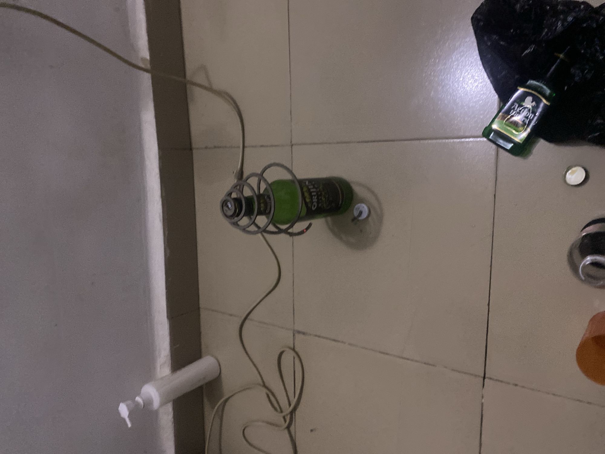 The image depicts a close-up, slightly blurry view of a tiled floor in a room, likely a residential setting in Howayi, Nigeria. The lighting is dim, creating shadows and highlighting the textures of the floor tiles and the objects present.
Dominating the center of the frame is a green glass bottle, possibly containing a beverage, with a metallic coil wrapped around its neck. A thin, light-colored cable is connected to this coil and extends out of view. Next to the bottle, on the tiled floor, lies a small, circular cap. To the left of the bottle, a white pump dispenser bottle is positioned near a wall. The wall appears to be a light color, and the floor is made of square, beige tiles. In the bottom right corner, a small green bottle with a label is partially visible, placed next to a crumpled black plastic bag. Other unidentifiable objects are also present at the bottom of the image. The overall impression is one of a domestic space with items scattered on the floor.