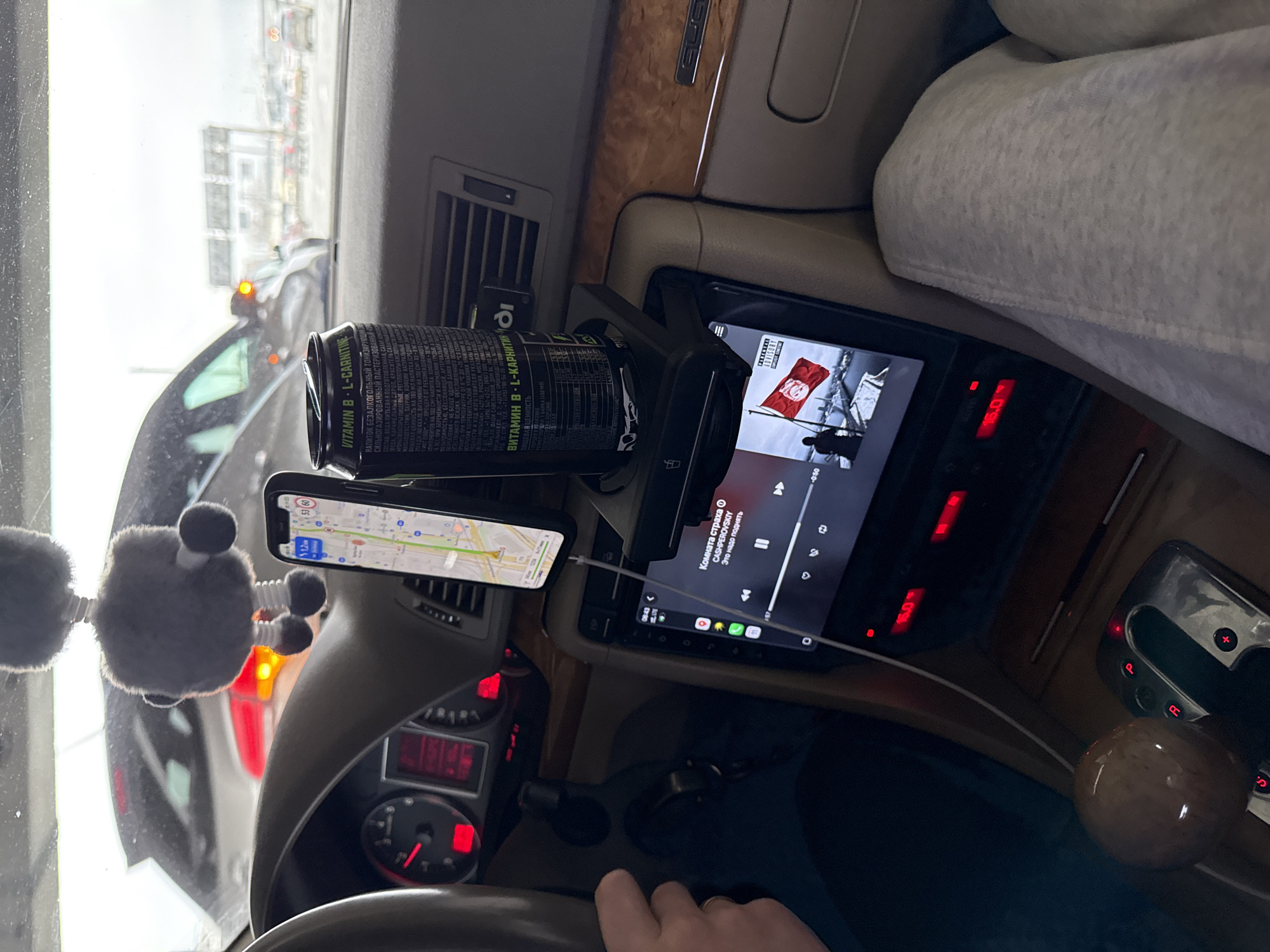 The image shows the interior of a car, likely an Audi given the emblem on the dashboard. The location appears to be a street in Moscow, Russia, based on the song playing on the in-car tablet by Russian artist CASHPEROVSKIY. The car's infotainment system displays the song "Комната страха" (Room of Horror), with album art featuring what seems to be a war memorial and the flag of Tunisia. The car is stationary, as indicated by the red "P" on the gear shift. The temperature display on the dash shows 16 degrees Celsius.
A smartphone is mounted to the dashboard, displaying a navigation map, likely used for directions. Next to it, a can of an energy drink (containing Vitamin B and L-Carnitine with Russian text) is held in a drink holder. A fuzzy car accessory is attached to the windshield, obscuring the view. A hand is visible holding the steering wheel.