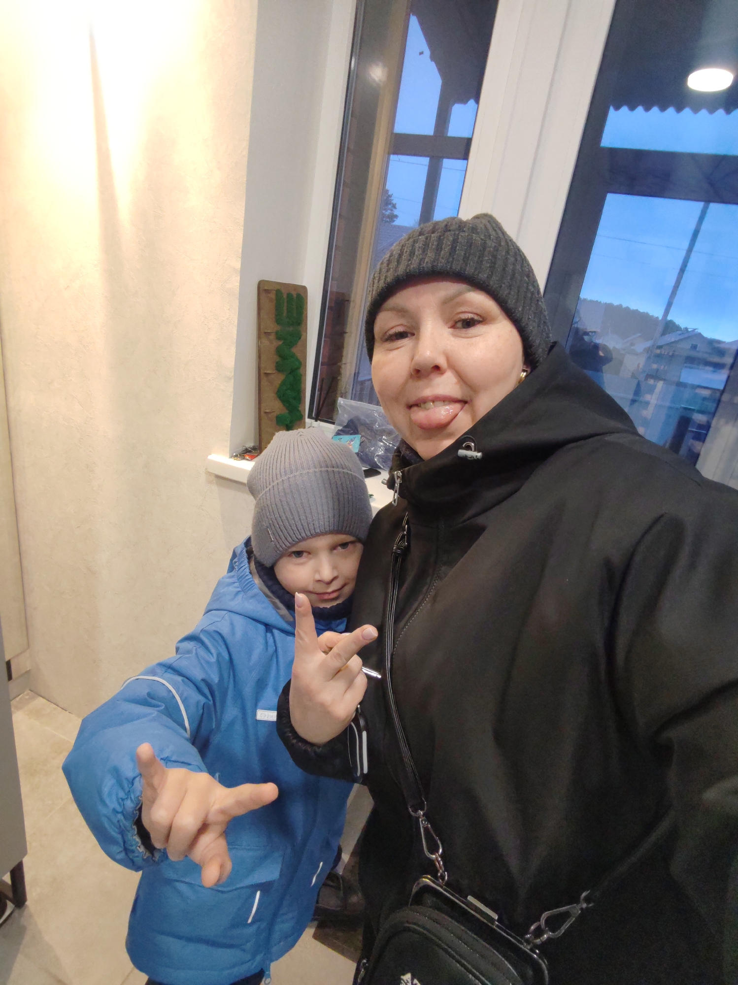 Two individuals are posing for a photograph indoors, with a window showing the outside in the background. The person in the foreground is wearing a black winter coat and a dark grey beanie, with a small black purse slung over their shoulder. They are sticking their tongue out and making a hand gesture. Behind them is a child wearing a blue winter coat and a light grey beanie, also making a hand gesture.
The window behind them reveals a blurred view of an exterior landscape, including buildings and what appears to be a hillside, suggesting an urban or semi-urban setting. The light suggests it might be dusk or early evening.
Near the window there is a wooden sign with green lettering, partly obscured. The interior wall is painted a light color. Without additional context, it is not possible to verify the location as Chaikovsky, Russia.