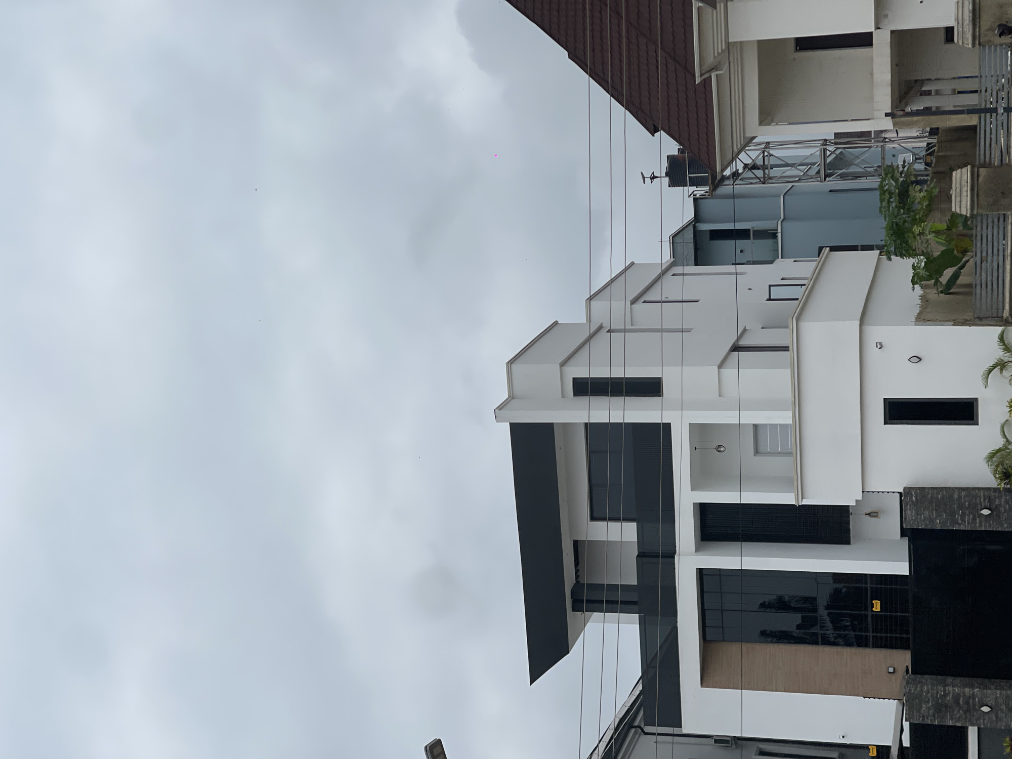 This is a ground-level, slightly low-angle, exterior, eye-level shot of several modern multi-story houses in Abuja, Nigeria. The sky is overcast with a light grey cloud cover. In the foreground, a tall black metal gate is visible, partially obscuring the lower parts of the houses.
The most prominent house, in the center and left of the frame, is a striking modern design with white and black facades. It features a large, dark tinted glass window that spans multiple stories on its left side, reflecting the surrounding environment. Above this, a section of the building is clad in a light brown material. A dark, angular overhang juts out from the top of this house. There are several black vertical window elements on the white sections.
To the right of this main house, another house is visible. It is predominantly white with a dark brown, sloped roof. The right edge of this house shows a white column and an open-sided ground floor. Further to the right, the very edge of another white building with a dark, narrow window is seen.
Behind the main house, a lighter blue house is partially visible, with dark windows. A metal structure, possibly an external elevator or a support for utilities, stands in front of the blue house. A black water tank with an antenna-like structure on top is situated on the roof of the blue house.
Electrical wires are strung across the middle of the frame, in front of the houses. A street light is visible on the far left, extending from the top left corner of the image. Some green foliage is present at the bottom of the frame, near the gate, suggesting some landscaping. The overall impression is of contemporary architecture in a residential area.