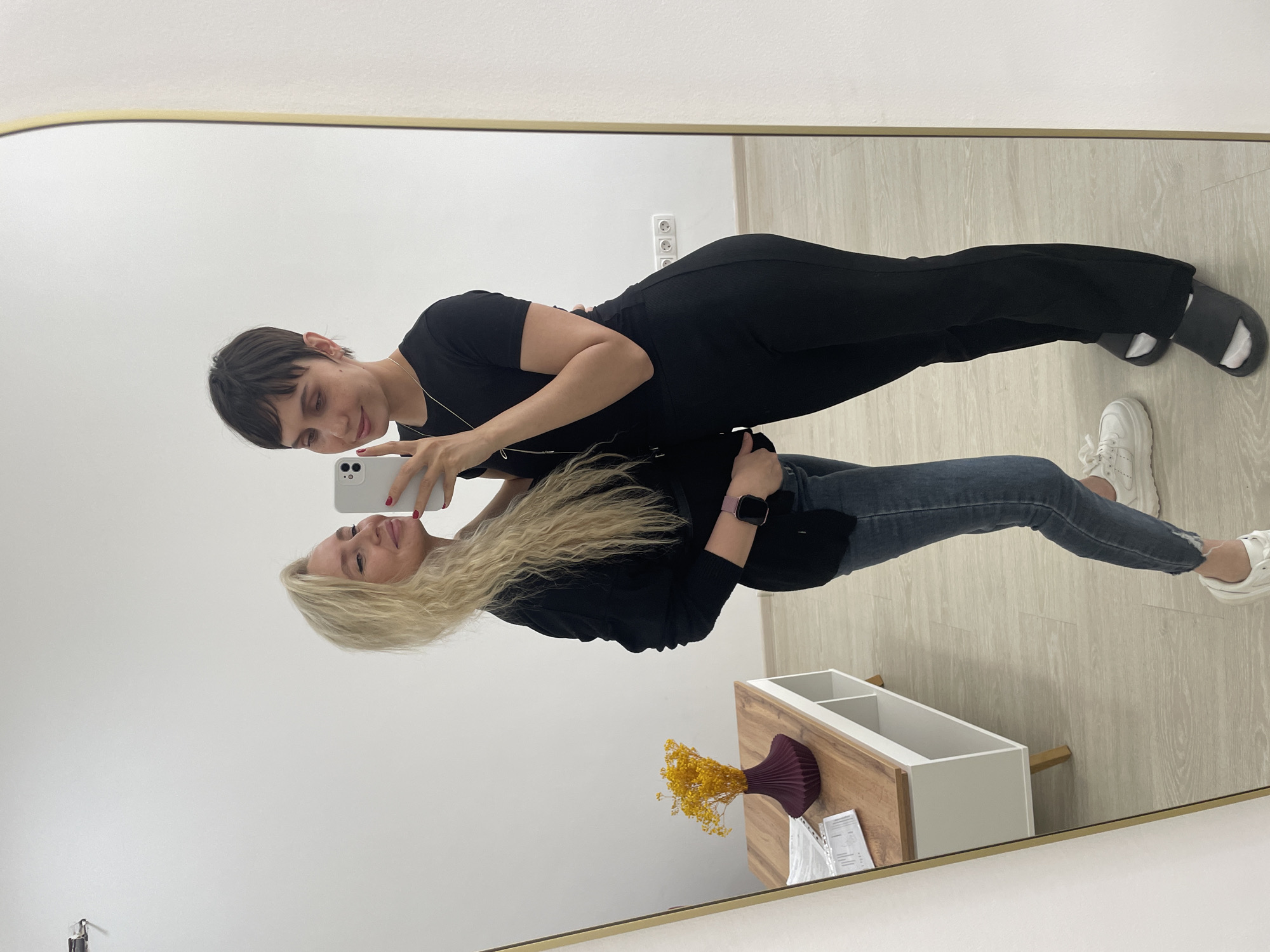 Two women are visible in the reflection of a full-length mirror. One woman is wearing a black t-shirt and black pants, and the other is wearing a black top and blue jeans. Both are looking at a white smartphone that the first woman is holding. The first woman has short, dark hair and a necklace. The second woman has long, blond hair. The scene appears to be indoors, with a white wall and light-colored flooring. There is a wooden console table with a vase of yellow flowers, and what appears to be a document on it. An electrical outlet is visible on the wall. Location is not specified.