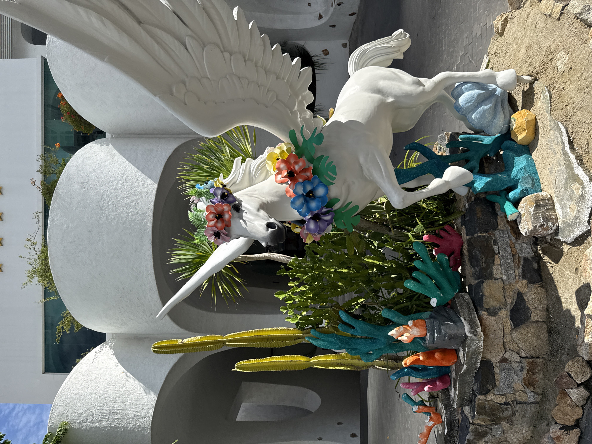 A white sculpture of a winged unicorn, adorned with colorful flower garlands around its neck and horn, stands prominently in an outdoor courtyard. The unicorn is positioned near a cluster of tall, yellow columnar cacti and lush green foliage, all set against a backdrop of a modern white building with curved architectural elements. The foreground features a decorative arrangement of colorful coral-like sculptures and rough-hewn stones. The location is identified as the area in front of the entrance to the Vinpearl Aquarium in Nha Trang, Vietnam, a popular tourist attraction situated on Hon Tre Island. The sky above is partly cloudy.