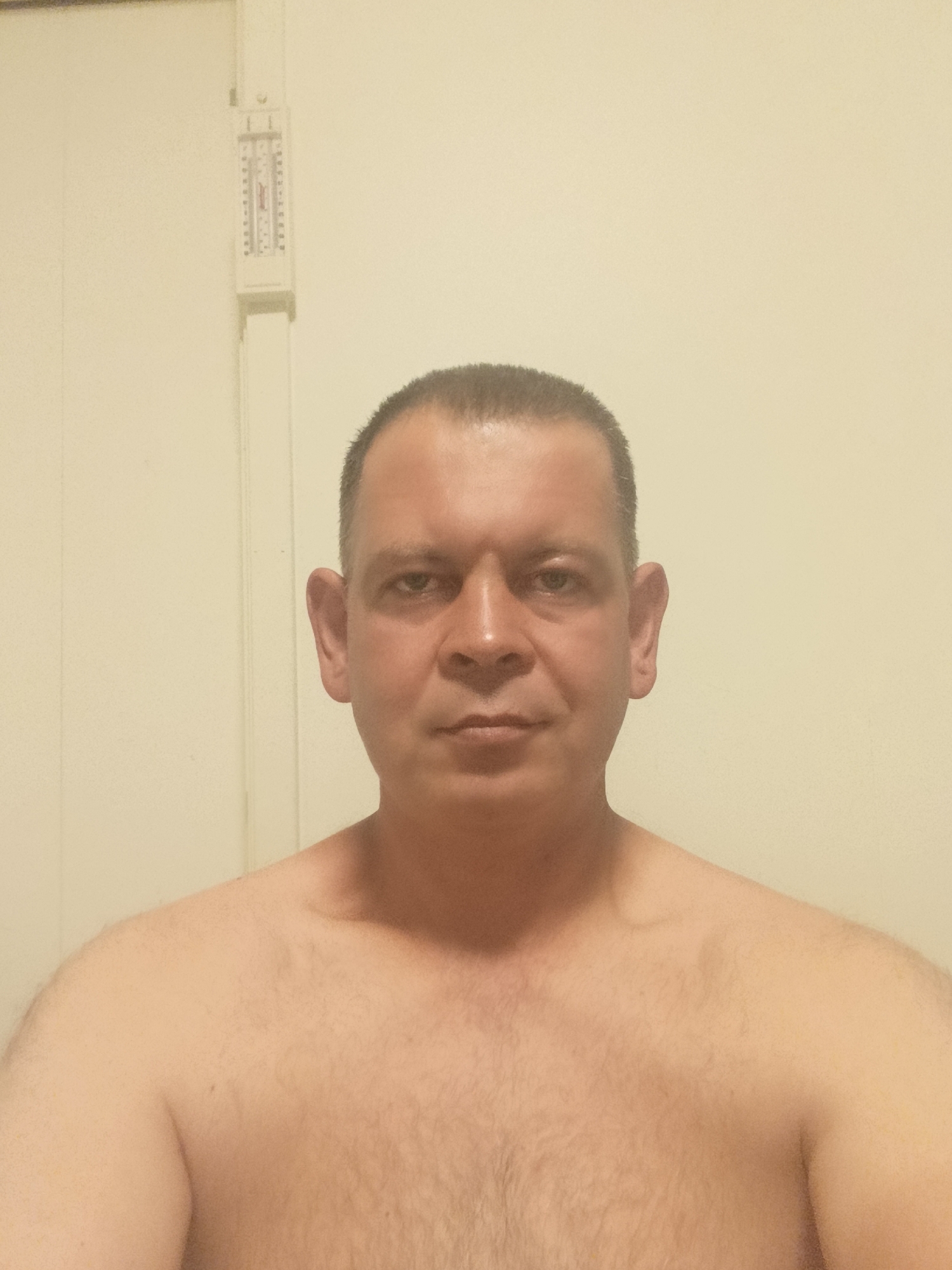The image shows a medium shot of a man standing shirtless in front of a pale yellow wall. He is facing the camera, with his head and upper torso visible. To the left of the man's head, on the wall, is a vertical white object with markings that resemble a thermometer or ruler. The man has short, dark hair, prominent ears, and a neutral facial expression. The lighting is somewhat dim and appears to come from the front. The background is a plain, slightly textured wall. There is no indication of a city or specific location, including Vallberga, Sweden.