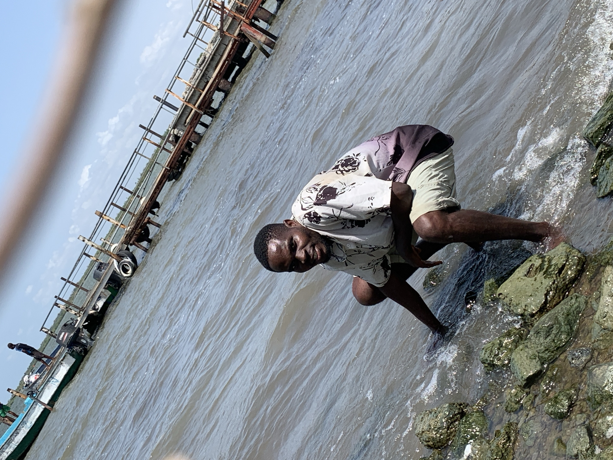 A man in a light-colored, short-sleeved shirt with a dark floral pattern and light-colored shorts is crouching by the water's edge on a rocky shoreline. He appears to be standing on some of the rocks, with his feet submerged in the water. The man is looking directly at the camera, and the photo seems to have been taken during the daytime.
The water is light brown and murky, with visible wave action near the shore. To the left of the man, there is a long, rusty structure extending over the water, supported by what appear to be railway car wheels. Further in the distance to the left, near the horizon, is a boat with a few individuals on board.
The location can be identified as Ikorodu, Nigeria due to the water and general environment.