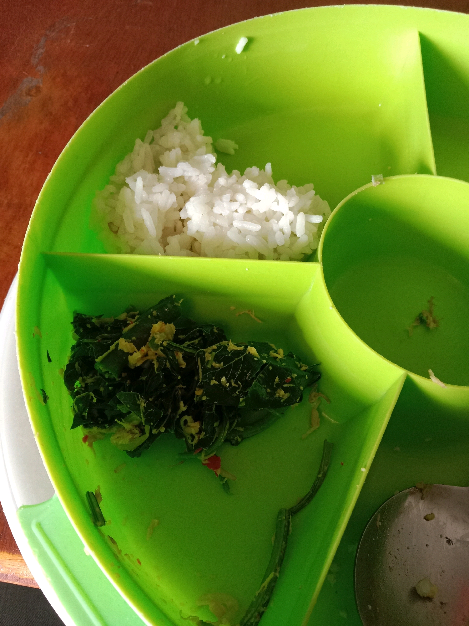 A close-up, high-angle shot shows a green, compartmentalized plate on a wooden surface, likely in Semarang, Indonesia. The plate contains white rice in the upper compartment and cooked dark green leafy vegetables with yellow specks in the lower left compartment. A round, deep compartment to the right is empty, with a small piece of food residue visible inside. A portion of a metallic spoon is present in the lower right section of the image. The lighting suggests an indoor setting, possibly a home or a local eatery. The wooden surface appears to be a table or countertop.