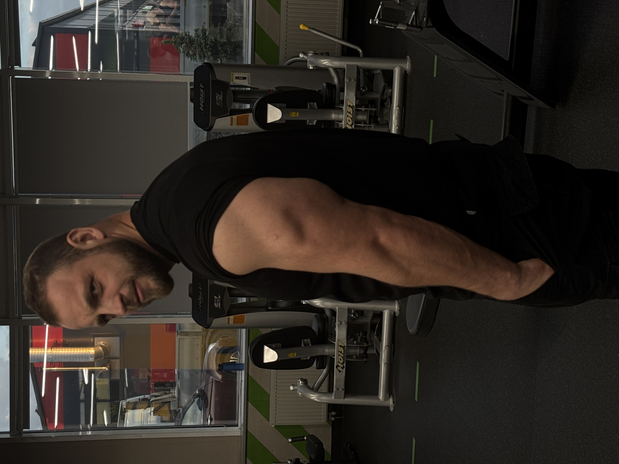 The image displays a man posing in an indoor gym setting. The subject, a man with short brown hair and a beard, is captured from the chest up, positioned horizontally due to the image's orientation. He wears a black short-sleeved t-shirt and dark pants, with his right arm flexed to emphasize his bicep and forearm. His right hand is partially obscured, appearing to be in his pocket or resting on his hip. He looks directly towards the viewer with a neutral, focused expression.
In the background, several pieces of gym equipment are visible, specifically grey and black strength training machines. The brand name "HOIST SL" and "HOIST" are clearly discernible on these machines. A large window stretches across the upper half of the background, offering a glimpse of an outdoor building with red and grey panels and some green foliage, possibly a commercial or urban structure. Reflections of vertical white indoor lights are prominent on the window. A white radiator is partially seen below the window, and the dark gym floor features subtle green line markings. The man's pose suggests he is showcasing his musculature, likely during or after a workout session. A small "DKNY" logo is faintly visible on his shorts in one of the cropped hints. The exact time of day is ambiguous due to the blend of artificial indoor lighting and visible external reflections.