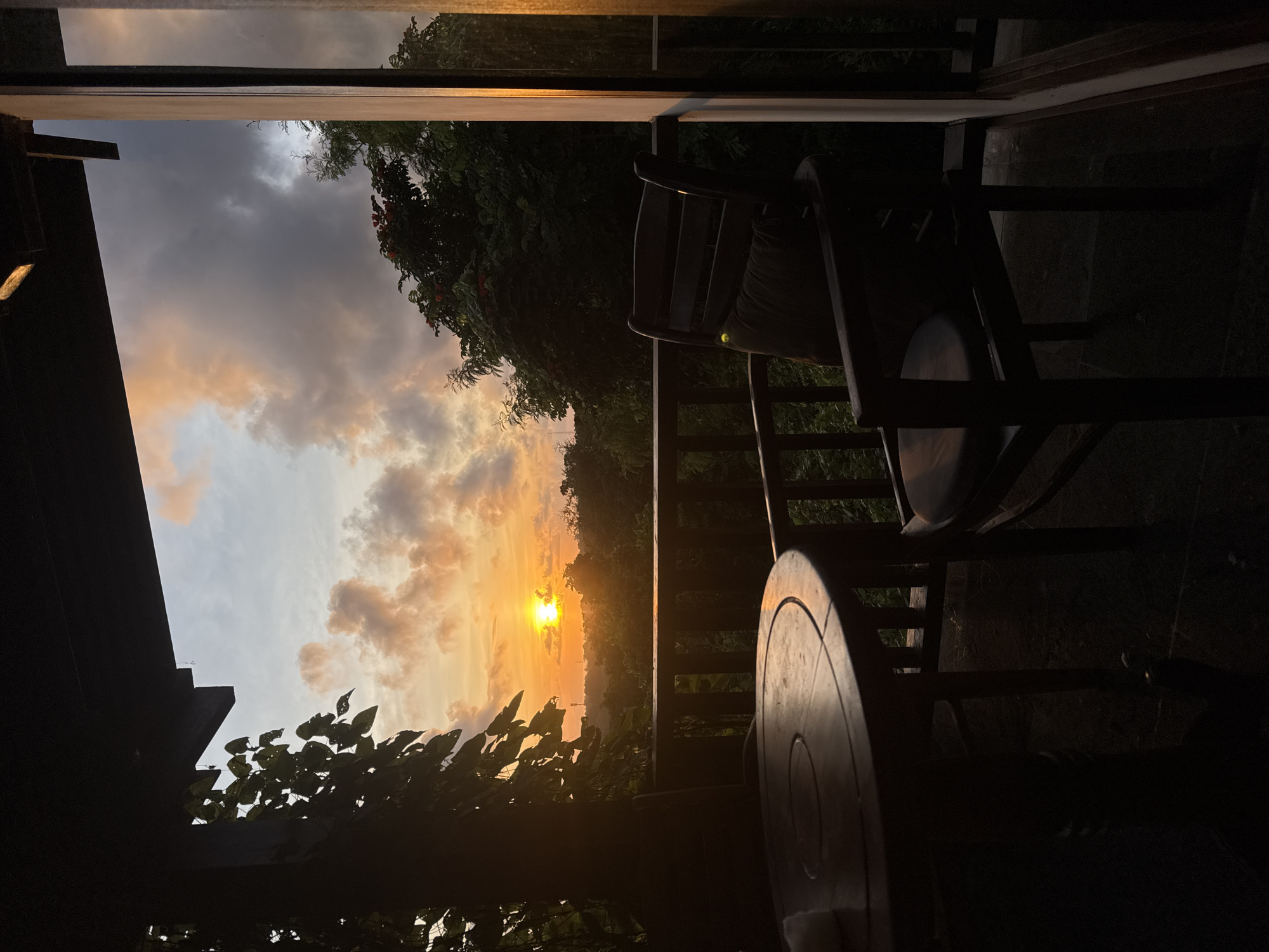 The image captures a sunset scene, possibly in Ungasan, Indonesia, viewed from what appears to be a balcony or veranda. Two dark-colored wooden chairs and a round table are silhouetted against the bright, orange-hued sunset. The sunset itself is partially obscured by trees and foliage, with the sun visible as a bright spot near the horizon. The sky above the sunset is a mix of orange, yellow, and gray clouds. The surrounding environment is mostly dark, suggesting the sunset is in progress. Part of a building roof is visible on the left. A handrail is visible, separating the foreground chairs from the view.