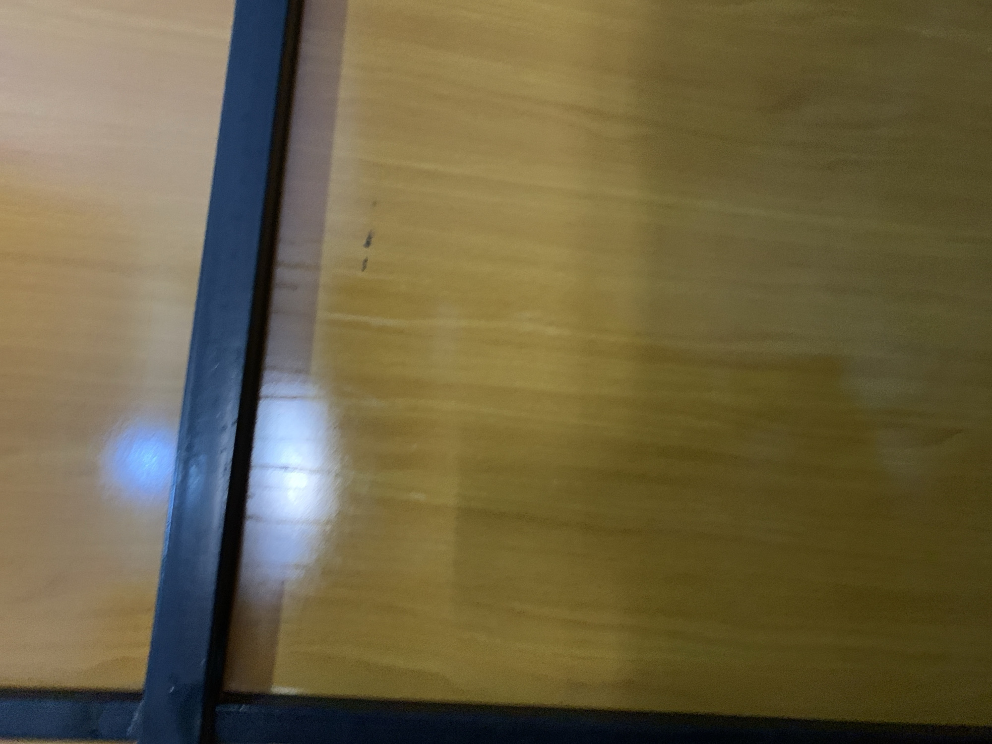The image shows a close-up, low-angle shot of a wooden surface, likely a door or paneling, with a dark horizontal bar running across the upper portion. The wood has a glossy finish, reflecting light in a bright, hazy blue oval in the upper center. There are subtle vertical wood grain patterns visible throughout the surface. A dark, vertical edge is present on the left side of the frame, suggesting this is a corner or the edge of a panel. A few small dark marks are scattered across the wooden surface. The overall lighting is dim, with the reflections from the light source being the brightest elements. The scene appears to be indoors, as indicated by the reflective and finished nature of the material. No people or specific locations within a city are identifiable.