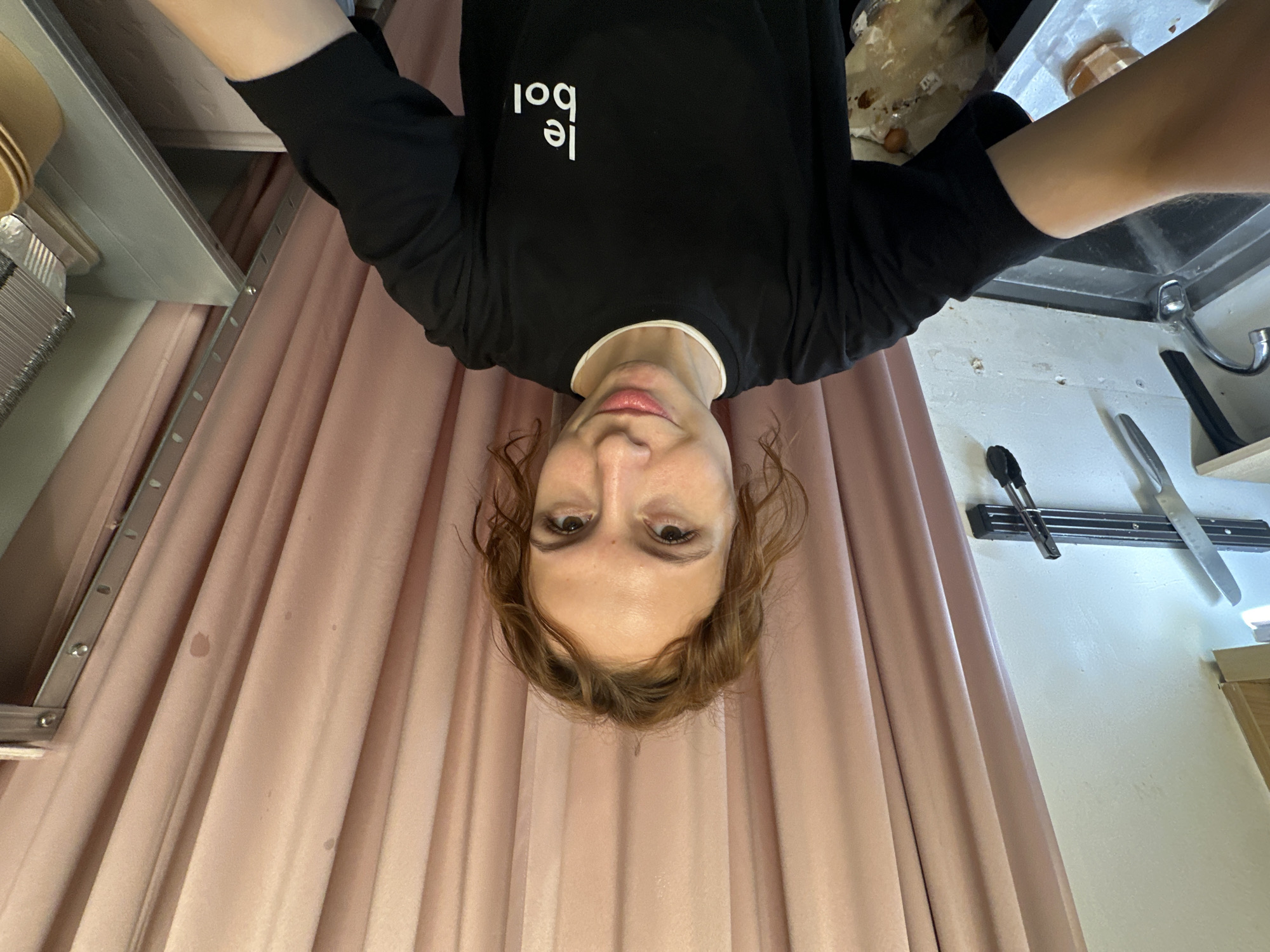 This inverted full-shot image features a young adult, likely female, with light brown, curly hair and a neutral expression, looking directly downwards towards the camera. Their face, with brown eyes and light pink lips, is positioned at the bottom center of the frame. They are wearing a black long-sleeved shirt with a white collar trim, on which the text "le bol" is printed in white, sans-serif letters, appearing upside down. Their arms extend upwards and are partially visible.
The background is dominated by light pink or peach pleated fabric, possibly curtains, running vertically behind the subject. On the left side of the frame, a grey shelf or countertop holds stacked beige bowls or containers, with what appears to be aluminum foil trays below them. A metal rail or track with small, circular fastenings is visible along the upper edge of the curtains on this side.
To the right, a stainless steel sink with a faucet is partly visible, suggesting a kitchen or food preparation area. Above the sink, a light-colored wall displays a black magnetic strip holding a large knife and a pair of tongs. The wall itself has several small, dark marks.
The scene suggests an indoor, functional setting, possibly a kitchen or food service environment in Novi Sad, Serbia, although no specific regional cues are present. The image captures a candid moment, likely a selfie taken from a low angle.