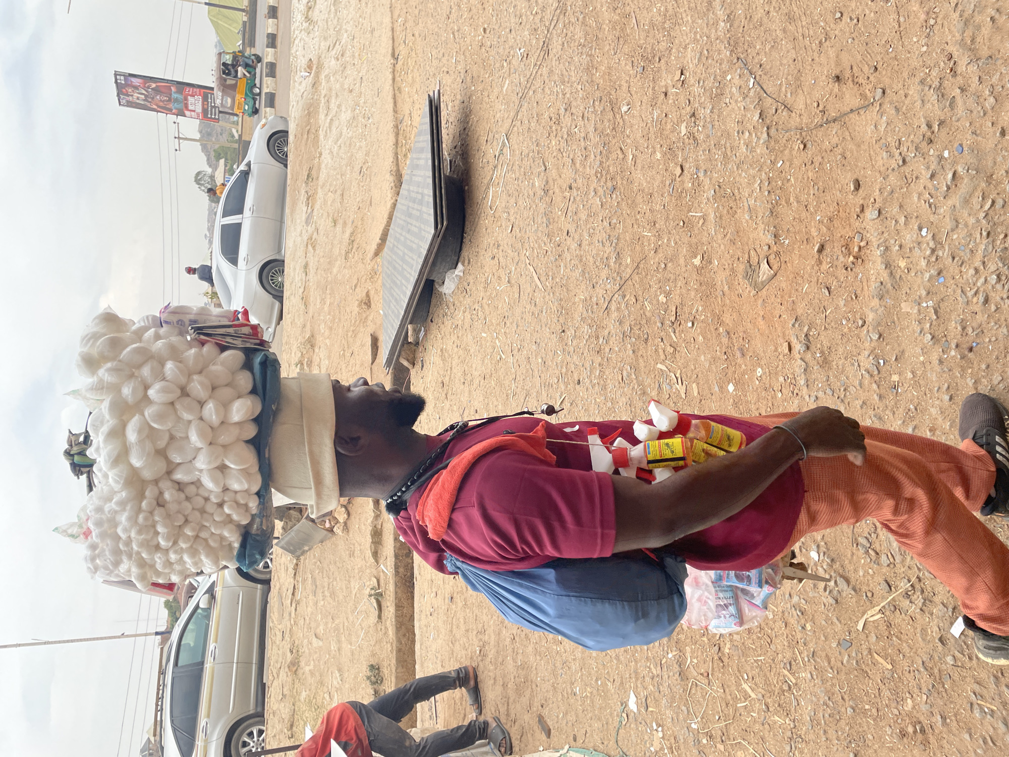 A male individual is depicted in a right-to-left stride on an unpaved reddish-brown dirt path in Jos, Nigeria. He is dark-skinned with a beard, wearing a maroon short-sleeved shirt, orange patterned trousers, black athletic shoes, and a light-colored cap. A large, white, irregular bundle, composed of numerous ovate white objects, is balanced on his head, supported by a blue cloth pad. He carries a blue drawstring backpack, and an orange cloth is draped over his left shoulder. Around his chest, multiple small spray bottles and clear plastic bags with contents are suspended by straps.
In the background, a white Lexus RX SUV is visible on the left, parked off the road. Further back, a silver sedan is present, with a person seated on its trunk. Beyond this, a green and yellow three-wheeled vehicle (keke napep) is seen. A large billboard with the text "INSTANT MIRACLES" stands prominently against a cloudy sky and distant hilly terrain. To the right of the main subject, a stack of four dark rectangular panels rests on two black cylindrical supports on the dirt ground. The immediate foreground and midground consist of dry, unpaved reddish-brown earth with scattered small rocks and debris. A second individual in a red shirt and dark trousers is partially visible, bent over, in the far left foreground.