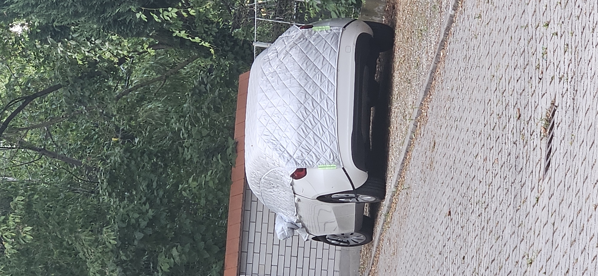 An unoccupied, white car is parked on a paved surface that appears to be a driveway or parking area. The car is partially covered by a quilted, silver-colored car cover, which extends over the roof and the rear of the vehicle. The cover has diamond-shaped stitching and some green accents are visible near the taillights. The car is positioned adjacent to a brick wall with a terracotta-colored roofline. Behind the car and wall, dense green foliage forms a natural backdrop. A metal fence is partially visible to the right of the frame. The ground in the foreground is paved with interlocking rectangular stones, with some sparse vegetation and leaves scattered on the surface. A drain grate is visible in the paved area. The scene appears to be outdoors, likely in a residential or private area in Hévíz, Hungary.