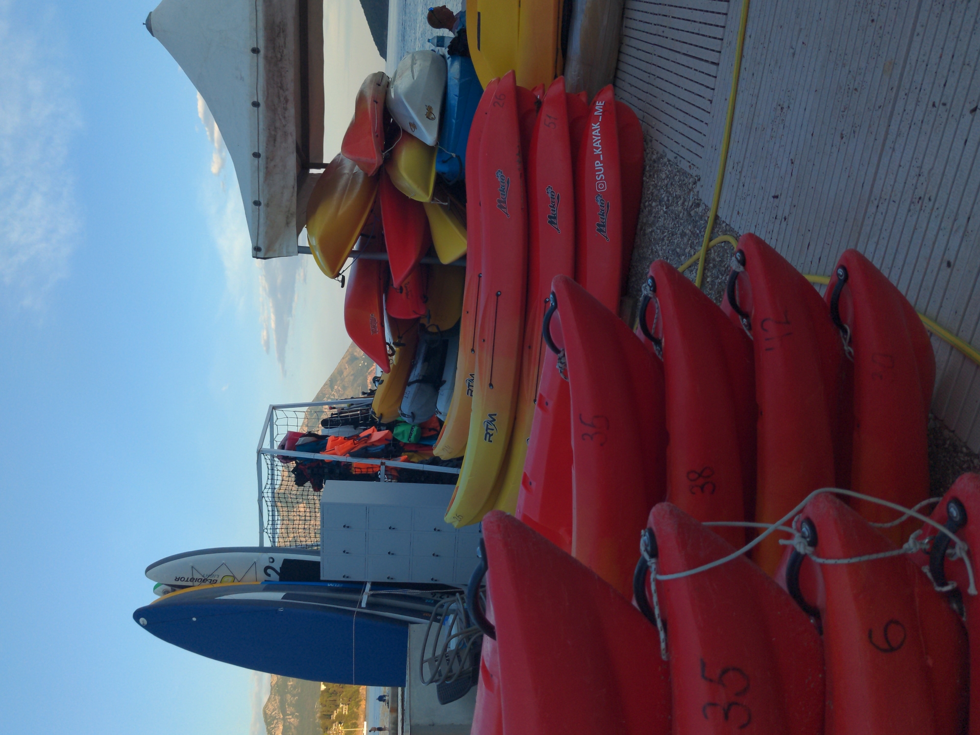 On an outdoor paved area in an unidentified city, numerous kayaks, mostly red and yellow, are stored on tiered racks. Life jackets and other equipment are in a caged section of the rack. Paddleboards are stacked near the kayaks. The sky is visible in the background.