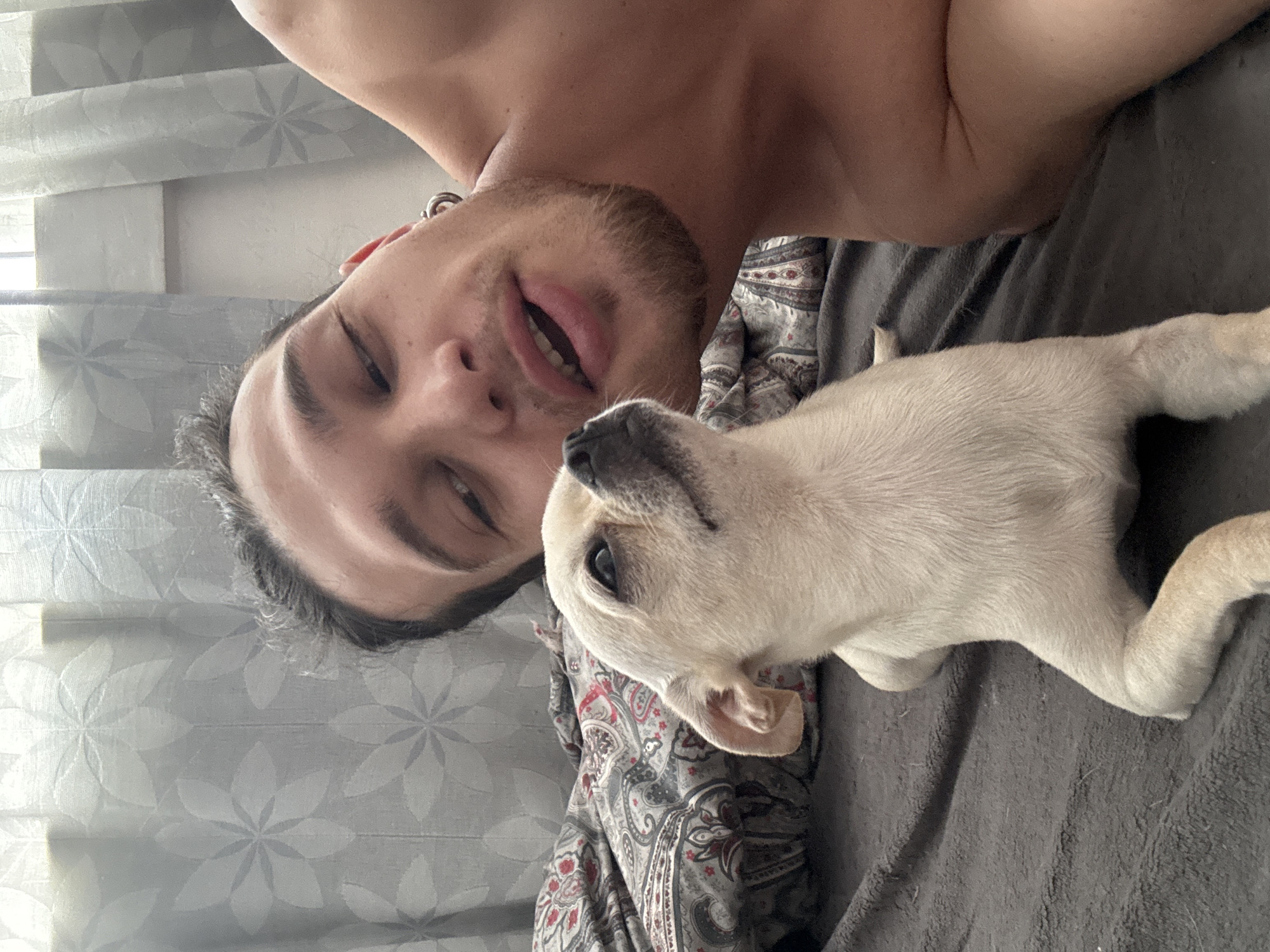 A man and a tan-colored puppy are in an indoor location. The man is light-skinned with dark hair and a beard, and is shirtless. He is looking at the camera with his mouth open. The puppy is lying next to him, looking up. The man is lying on a bed with a paisley-patterned comforter. Behind him is a gray curtain with a floral pattern. The exact location cannot be determined from the image. There is no definitive indication that this photo was taken in Sofia, Bulgaria.