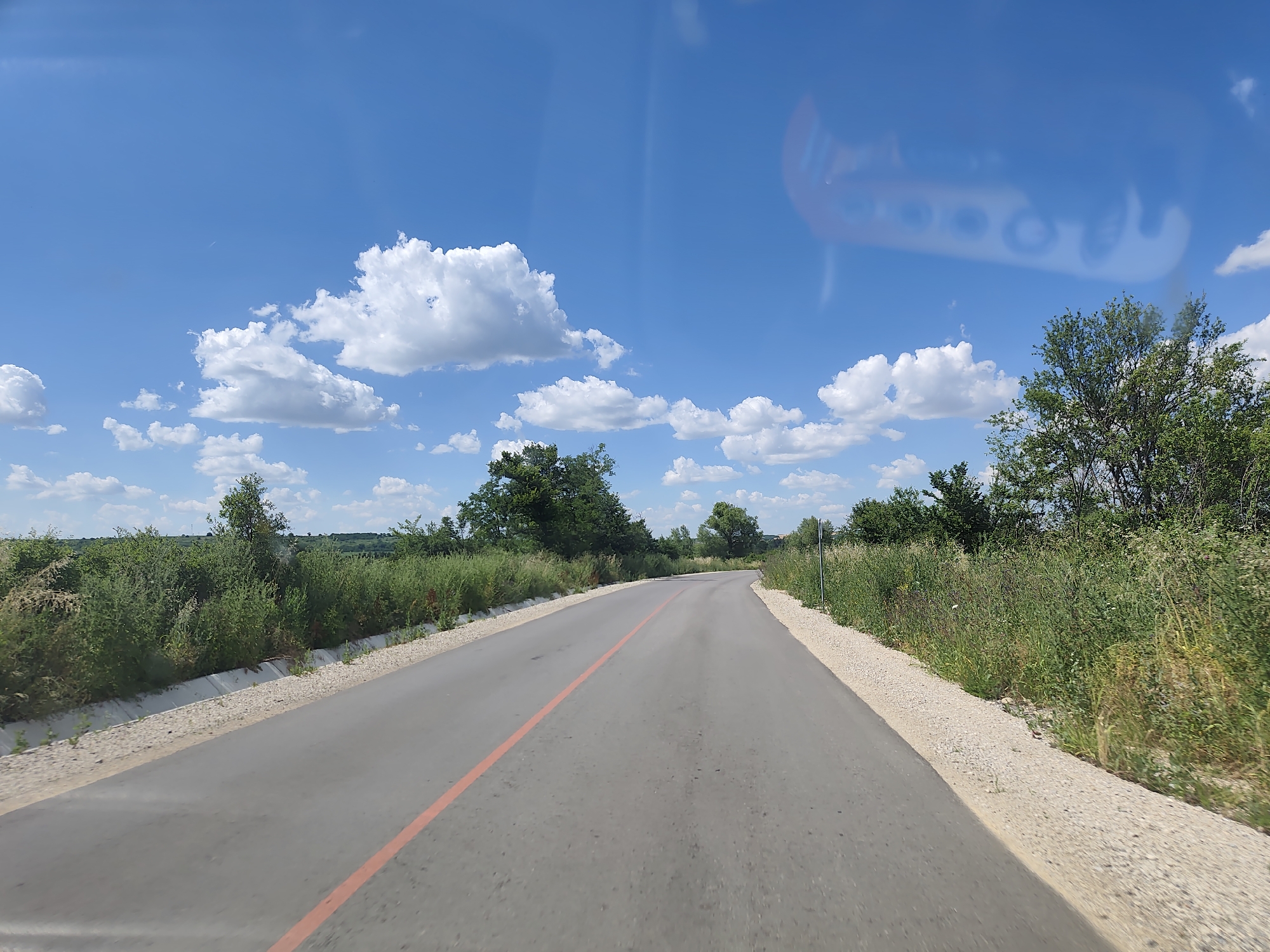 This media file captures a view from a vehicle traveling on a paved road through a rural landscape. The road, with a single orange line marking its center, curves gently towards the horizon. On either side of the road, lush green vegetation, including bushes and trees, lines the way. The ground beside the road is covered with gravel and some small drainage ditches. In the distance, rolling hills are visible under a bright blue sky dotted with fluffy white clouds. The scene suggests a sunny day, likely in the late morning or early afternoon given the bright illumination. There are no people or distinct objects visible in the scene, only the natural surroundings and the infrastructure of the road. The overall mood is one of peaceful travel through the countryside. A faint reflection, possibly from the vehicle's interior, is visible in the upper portion of the image. The location is identified as Dermantsi, Bulgaria.