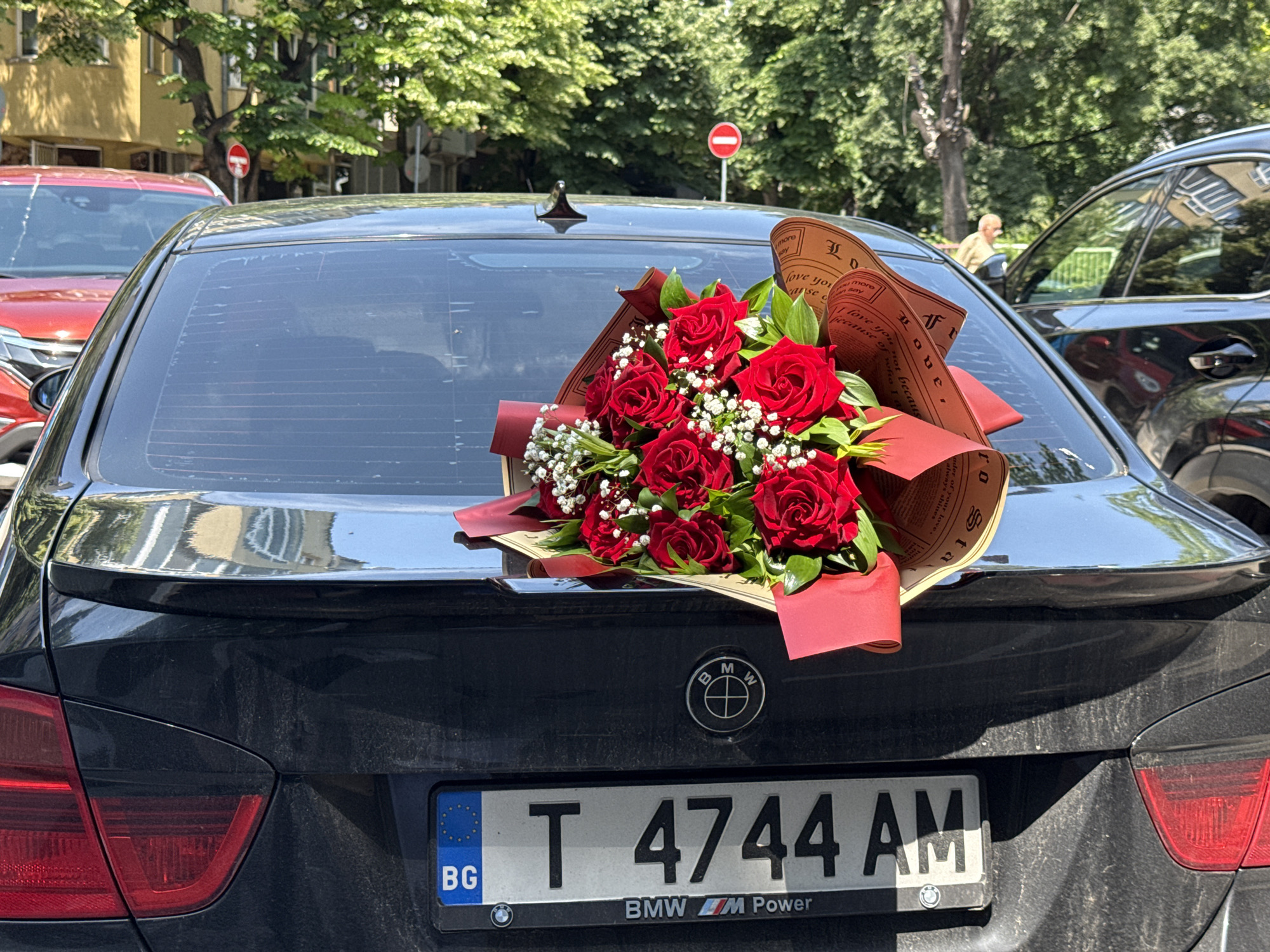 A black BMW car, likely a sedan, is prominently featured from its rear in bright daylight, parked on what appears to be a city street in Varna, Bulgaria. A vibrant bouquet of red roses, interspersed with baby's breath and green foliage, rests on the car's trunk, wrapped in reddish-brown paper adorned with text and a red ribbon. The car has a subtle black spoiler and a "BMW M Power" license plate frame. The Bulgarian license plate "BG T 4744 AM" is clearly visible. The car itself appears somewhat dusty.
In the background, lush green trees line the street, indicating a sunny day, likely during late spring or summer. To the left, part of a red car is visible, and to the right, another dark-colored vehicle is partially seen, reflecting nearby buildings. A blurred person can be discerned walking in the distance between the vehicles on the right. A circular red street sign with a white horizontal bar, signifying "No Entry," stands out amidst the greenery.
The bouquet's wrapping paper features fragmented English text, including phrases like "I love you," "because of what I love you not because," "Love," and "always shines," suggesting a romantic or celebratory gesture. No specific activity is captured, but the presence of the bouquet implies a special occasion, transport, or delivery. The overall scene evokes a pleasant, warm atmosphere in an urban setting.