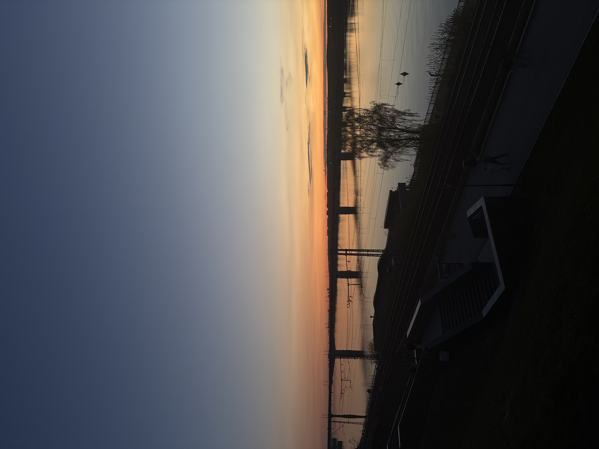 The image depicts a sunset over the Kama River in Perm, Russia. The sky transitions from dark blue to orange and yellow hues near the horizon. The river reflects the colors of the sunset, with the opposite shore appearing as a dark silhouette. Several power lines run across the river. A building and a tree are also visible in the foreground, with a pedestrian walking along a path. The lighting is dim, emphasizing the sunset and creating shadows in the foreground.