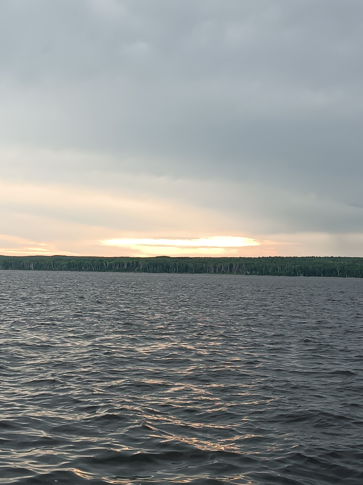 The image depicts a body of water, likely a lake or river, with a treeline visible in the distance under a cloudy sky. The water has small waves and reflects the muted light from the sky. The treeline is dense and appears to be a forest. The sky is overcast with a bright, illuminated band where the sun is likely breaking through the clouds. The scene is tranquil and appears to be taken from a boat or the shore. The location is identified as the Kama River near Chaikovsky, Russia.