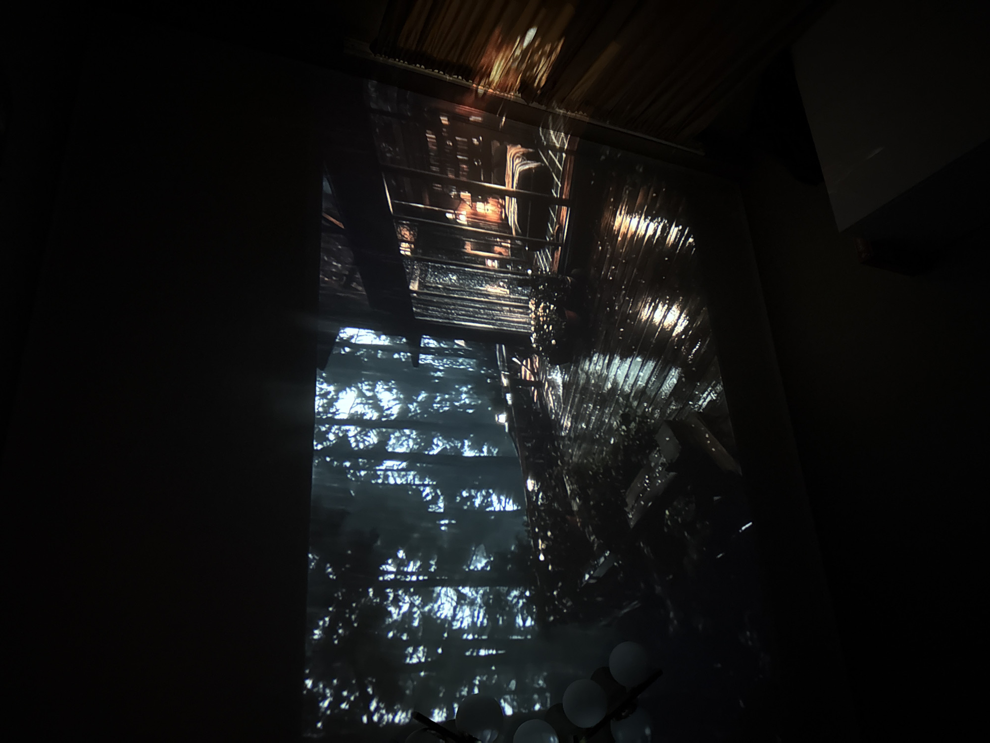The image shows a projection of a rainy, nighttime scene in a forest, with a modern cabin or house on the right. The cabin has large glass windows, through which a bedroom with a lit lamp and bookshelves is visible. The deck of the cabin is wet and reflects the lights. To the left of the cabin, there are tall, dark trees. In the foreground, on the far left, there are some round, illuminated objects that appear to be part of a light fixture. The overall ambiance is dark and moody, with a sense of being in a secluded, natural environment. There is no visible indication that this scene is located in Tbilisi, Georgia.