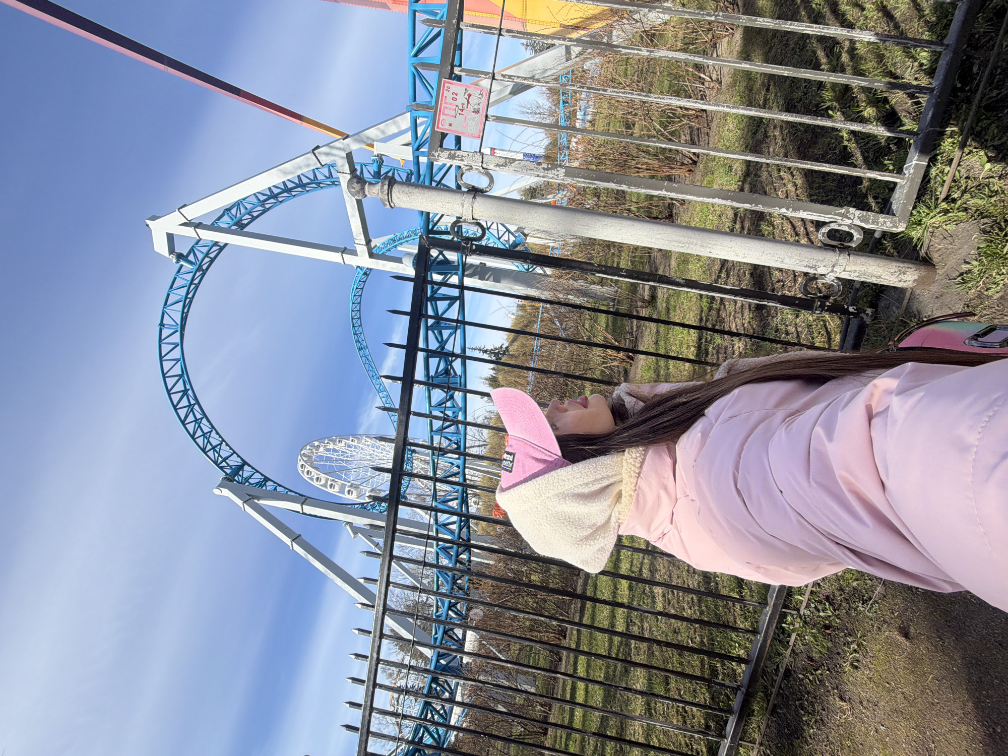 The image shows a young woman, wearing a pink hooded jacket and a pink hat, standing next to a black metal fence. Behind the fence are amusement park rides, including a roller coaster and a Ferris wheel. The sky is blue. The photo was taken at Divo Ostrov amusement park in Saint Petersburg, Russia.