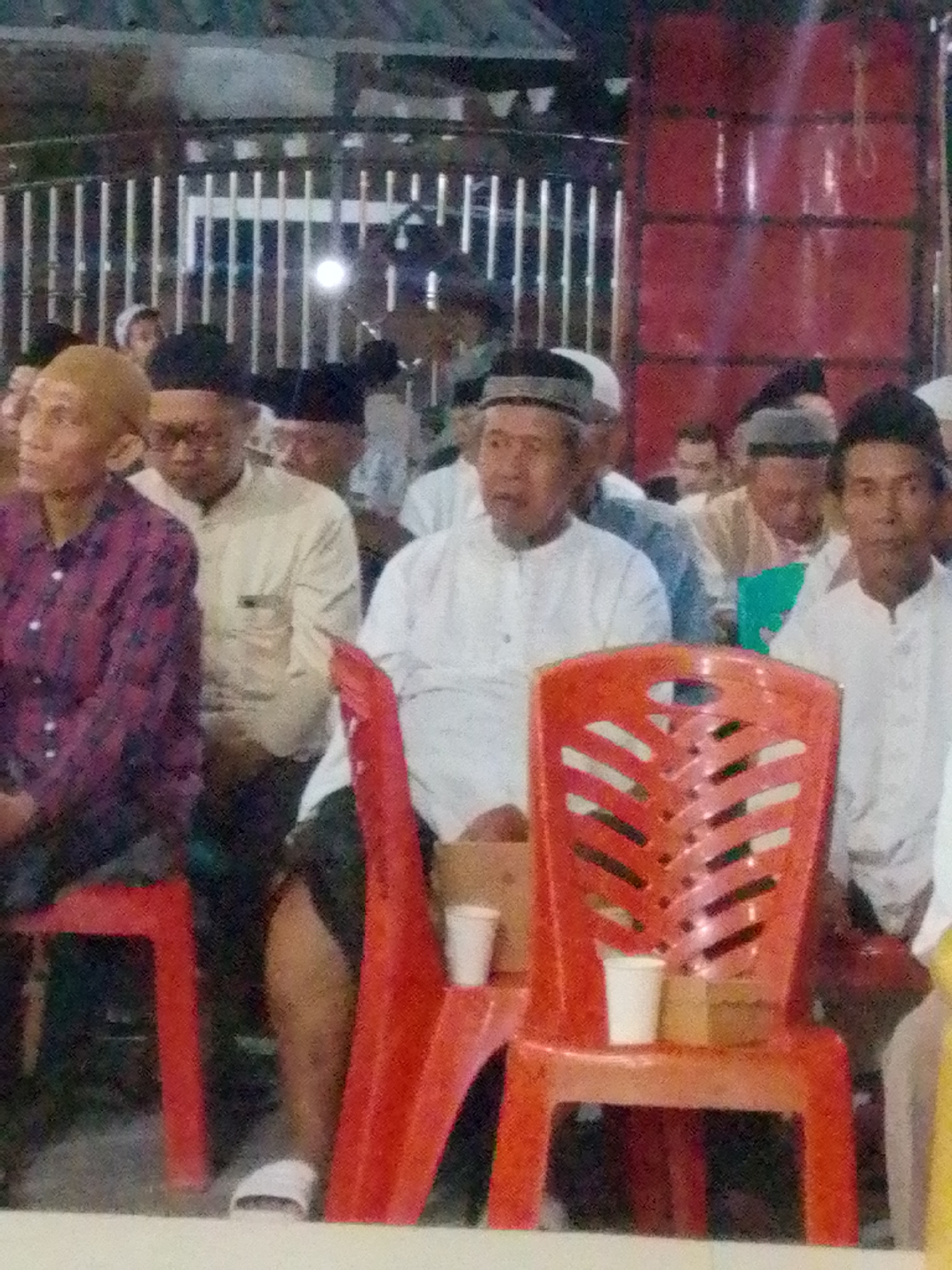 The image shows a group of men, likely gathered for an event, in an outdoor setting at night. The men are dressed in traditional attire, with some wearing skullcaps or turbans. The background features a fence and a red structure with horizontal divisions, possibly a building or stage. Red plastic chairs are visible in the foreground, with white paper cups placed on them. The overall lighting suggests artificial illumination. The scene appears to be from a public gathering in Bandar Lampung, Indonesia, likely an Islamic religious or community event.