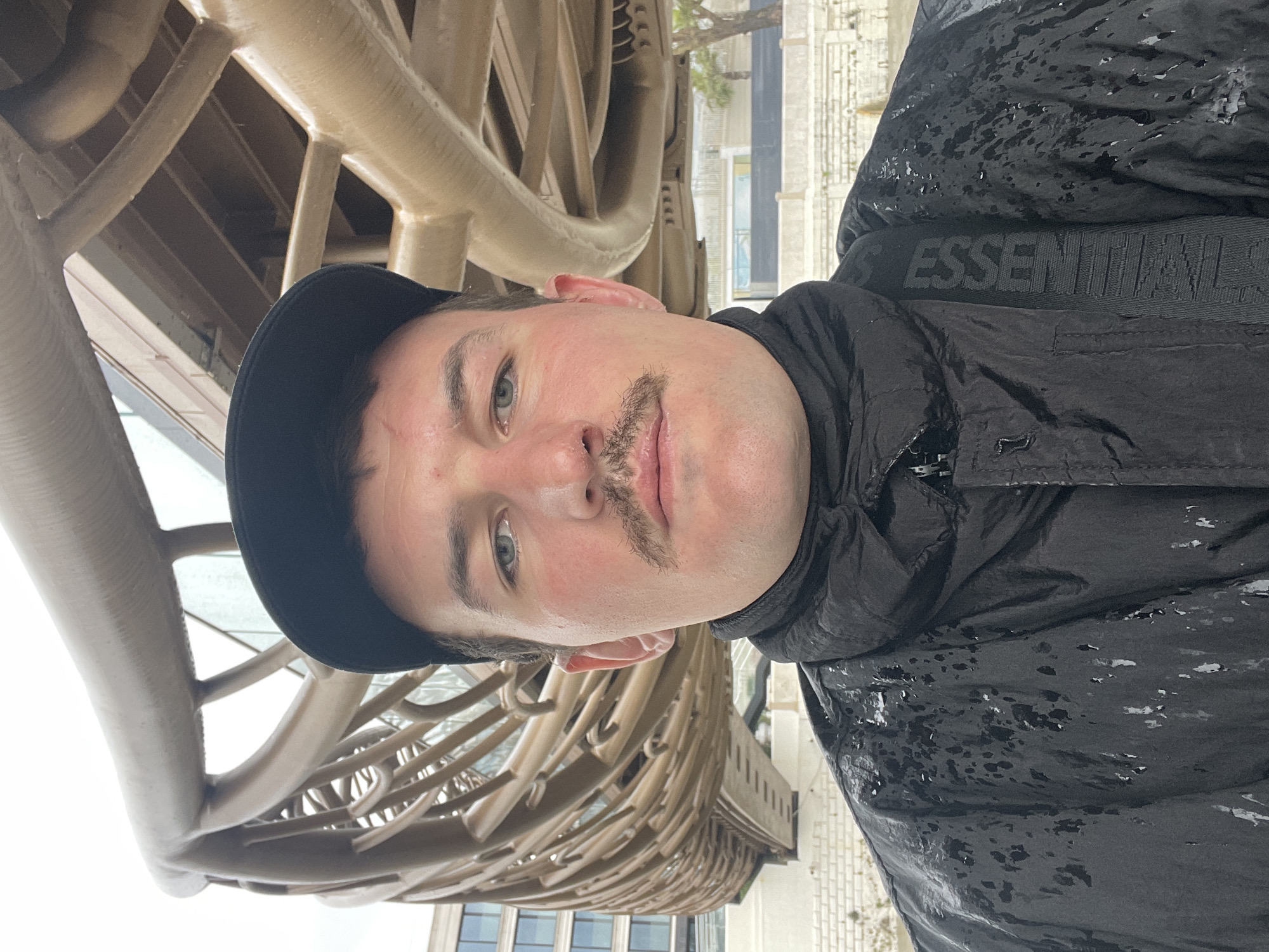 A man stands near the Sea Port of Sochi, Russia, with a section of the building visible in the background. The man is wearing a black cap and a black jacket with the word "ESSENTIALS" visible on the strap of a bag worn across his body. He has a mustache. The scene appears to be outdoors, possibly on a cloudy or overcast day.