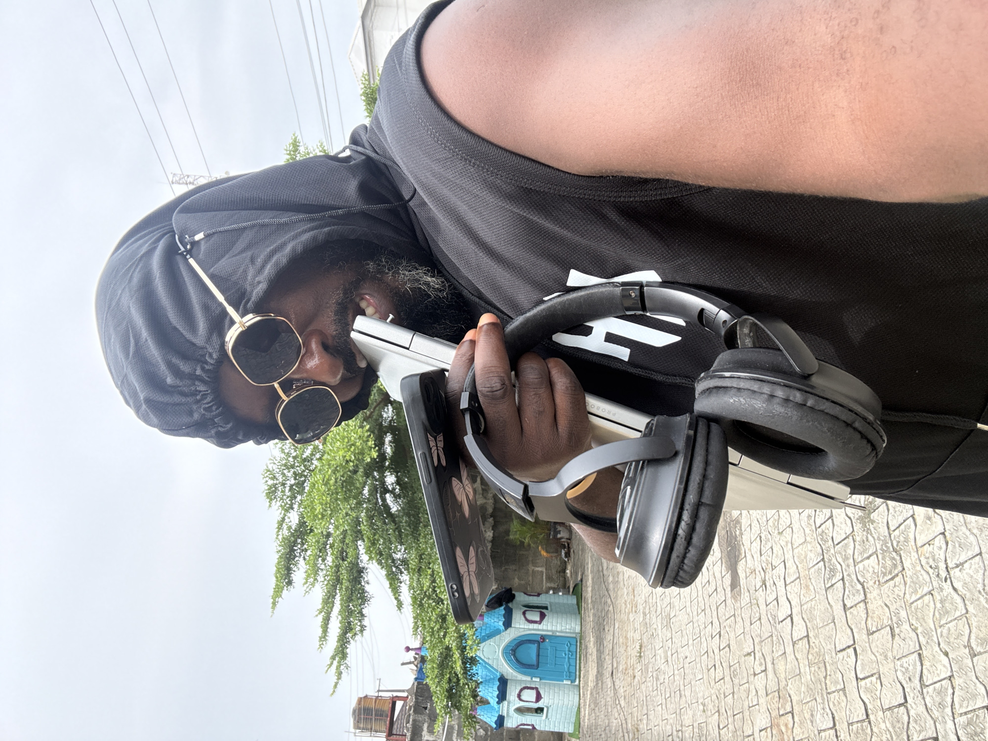 The image captures a man from a low-angle perspective, holding various electronic devices outdoors in Ado, Nigeria. He is seen from the chest up, wearing a black hooded top with the hood pulled over his head, and black sunglasses with golden-colored frames. A beard, containing some grey or white hairs, is visible around his mouth and chin. Underneath the hooded top, he wears a black sleeveless shirt featuring a partial white graphic or logo on its chest area.
The man holds a silver laptop in his right arm, with black over-ear headphones draped around it. In his left hand, he grips a black smartphone encased in a cover adorned with pink butterfly designs. His gaze is directed downwards, suggesting engagement with one of his devices.
The background shows an overcast, light grey sky and lush green trees. The ground beneath the subject is paved with light-colored, interlocking stones. In the mid-ground, a small, light blue and white plastic playhouse is visible. Further in the distance, partially obscured by foliage, a large structure, possibly a water tank on a stand, and other architectural elements can be seen. The scene is set during daytime under cloudy weather conditions. No distinct text is clearly visible on any of the objects or in the background, other than the partial white graphic on the man's shirt.