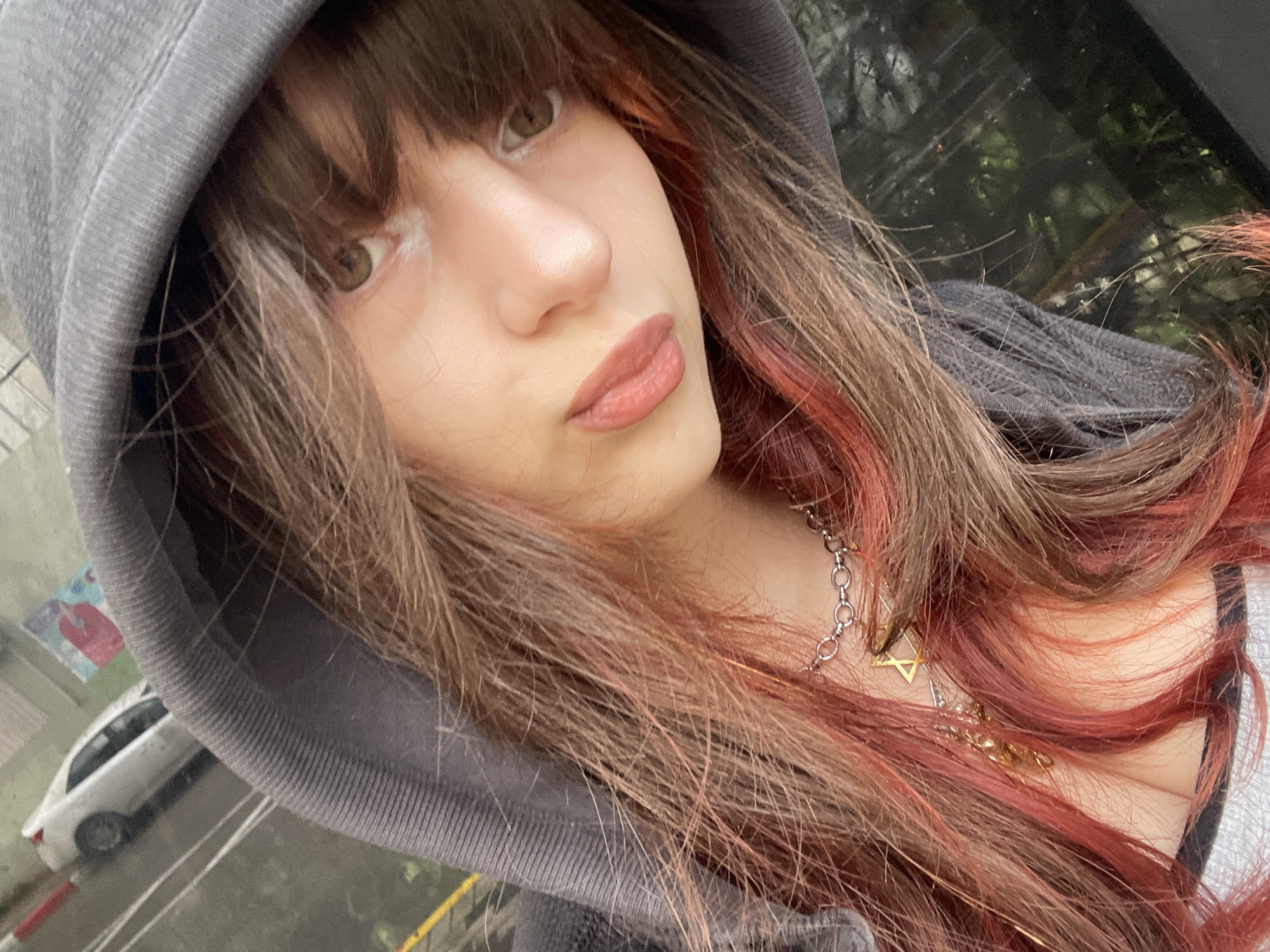 The image depicts a close-up, upward-angle portrait of an individual in Petah Tikva, Israel. The subject has long brown hair with prominent reddish-pink highlights, partially concealed by a grey hooded sweatshirt worn with the hood up. The face is visible, featuring large brown eyes, a small nose, and lips colored with a pinkish-red tone. Around the neck, a silver-toned chain necklace is worn, adorned with multiple pendants, including a geometric star-shaped pendant and another gold-colored pendant. A white garment with black stripes is partially visible below the necklace. The background is out of focus, revealing an outdoor urban environment under an overcast sky. To the upper left, a white sedan is visible on a wet asphalt road with yellow and white painted markings. A red pole and a faded, colorful sign are partially visible beyond the vehicle. To the right, green foliage is reflected in a dark, glossy surface, likely a window.