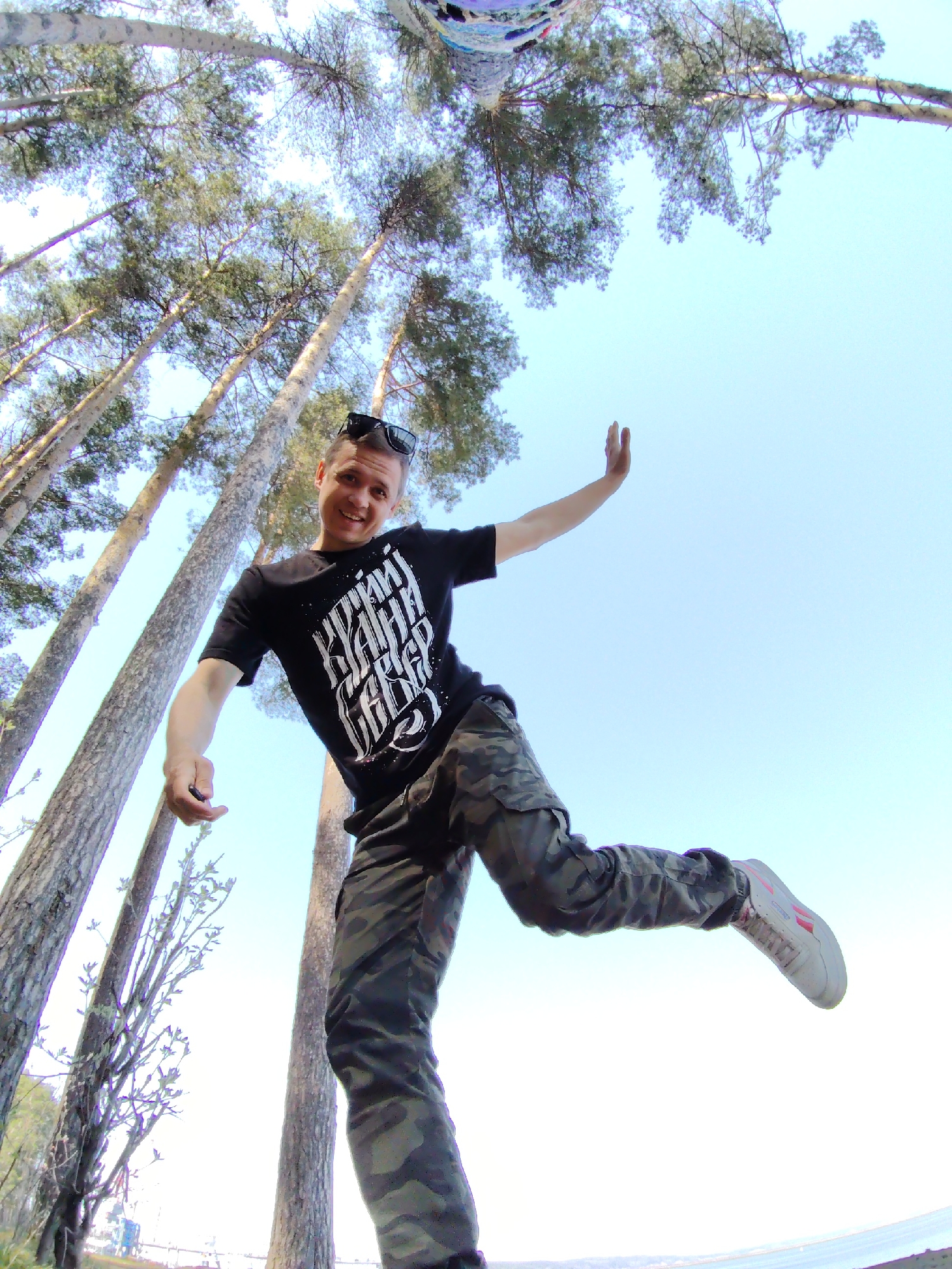 A man stands on one leg in an outdoor setting, possibly a park in Chaikovsky, Russia, surrounded by tall trees. The man is wearing a black t-shirt with white writing, camouflage pants, and tennis shoes. He is holding his other leg up in the air, and his arms are outstretched. He has sunglasses on his head and is smiling. The trees are tall and thin, and the sky is blue with some clouds. The photo appears to have been taken with a wide-angle lens, giving it a distorted perspective.
