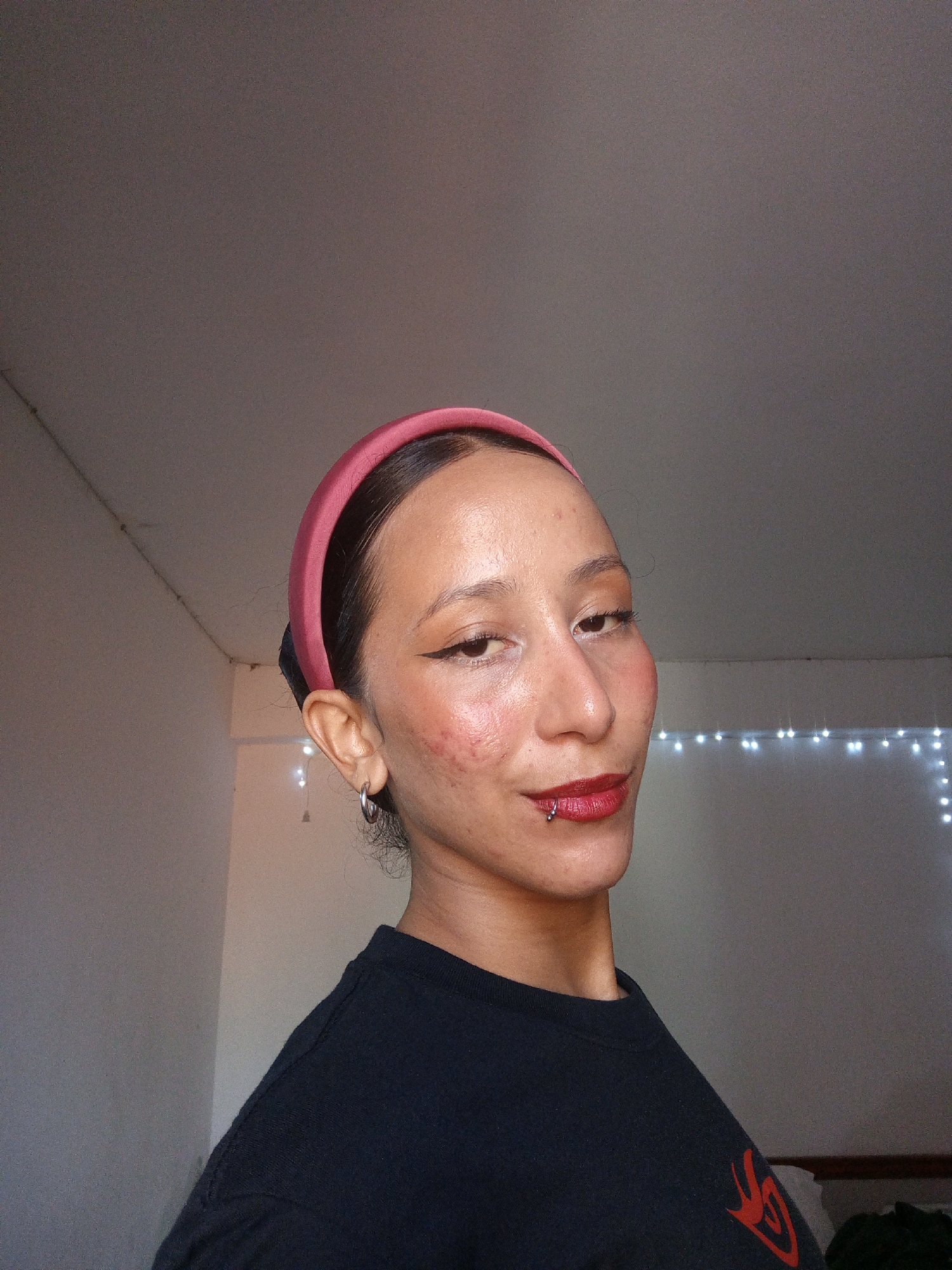 A person with fair skin, wearing a pink headband and dark lipstick, poses for a selfie. The person has a lip piercing on the left side of their lower lip. They wear a black t-shirt with a red design on the sleeve. Their left ear has a hoop earring. The background consists of a light-colored wall with a string of fairy lights. The exact location is unknown.