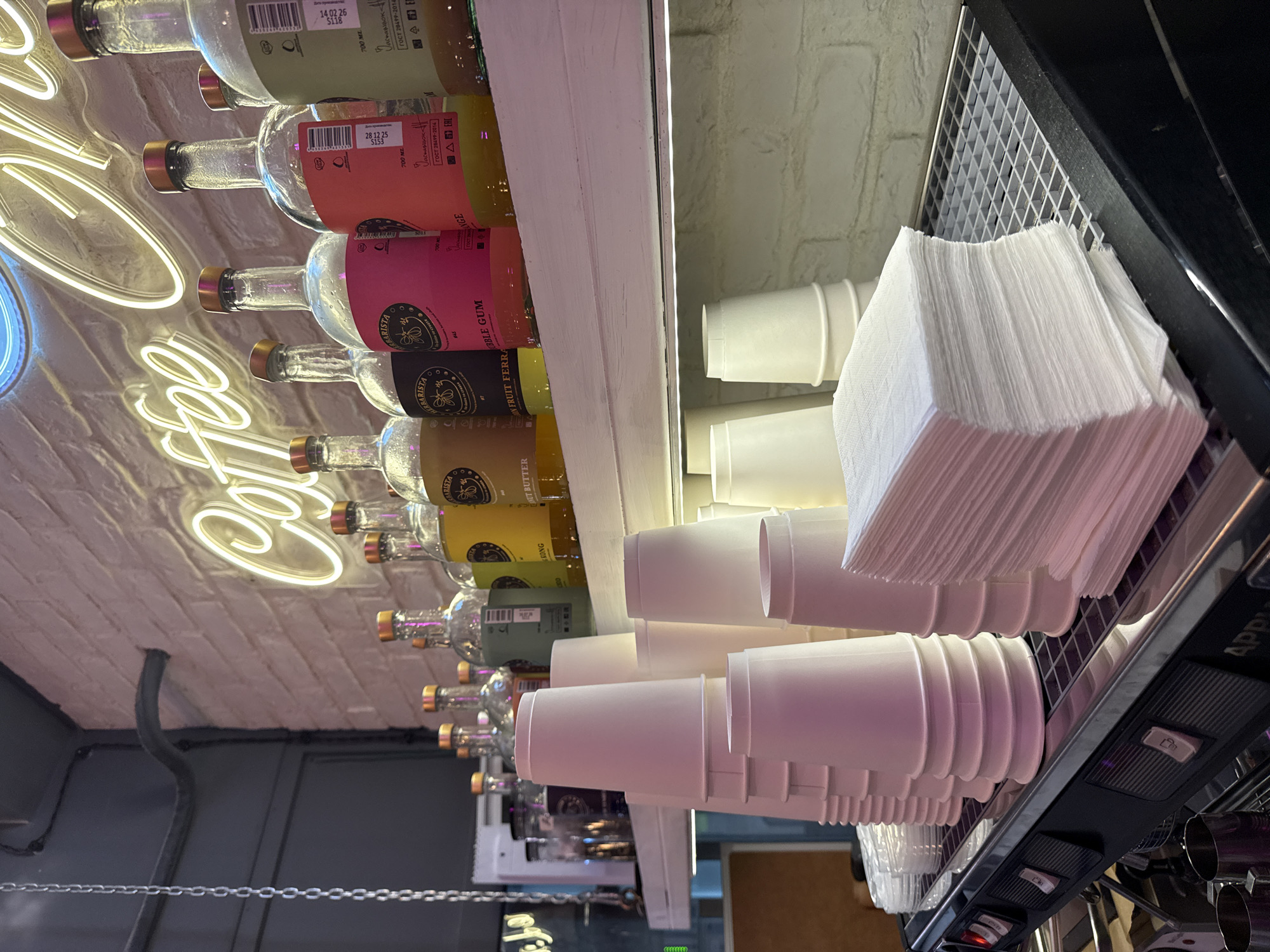 The image depicts a close-up, eye-level view of a coffee shop counter in Moscow, Russia. The scene is dominated by stacks of white paper cups on the left and a thick stack of white paper napkins on the right, both placed on a metal grate. Behind the cups and napkins, a white shelf displays a row of glass bottles with colorful liquids and gold caps. A neon sign, partially visible at the top, spells out "Coffee" in a cursive font. The background consists of a white brick wall. The lighting is artificial, creating a warm and inviting atmosphere. No people are visible in the image. The overall impression is one of a well-stocked and organized coffee preparation area.