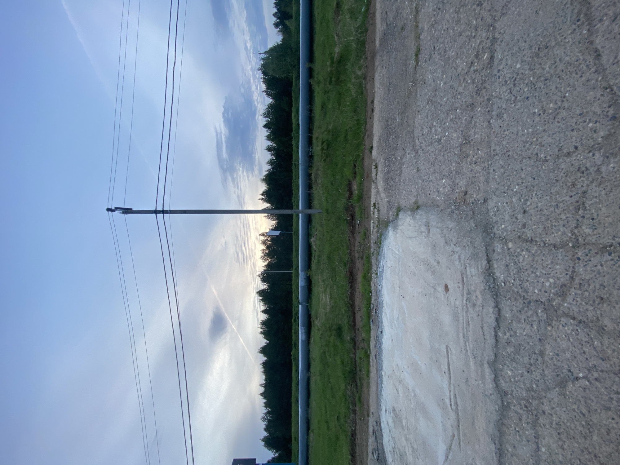 The image shows a view in Прикамский, Russia. An asphalt road runs along the right side of the image. To the left of the road is a grassy area bordered by trees and a long, dark gray pipeline supported by posts. Power lines stretch across the sky above the trees. The sky is light blue with streaks of clouds.