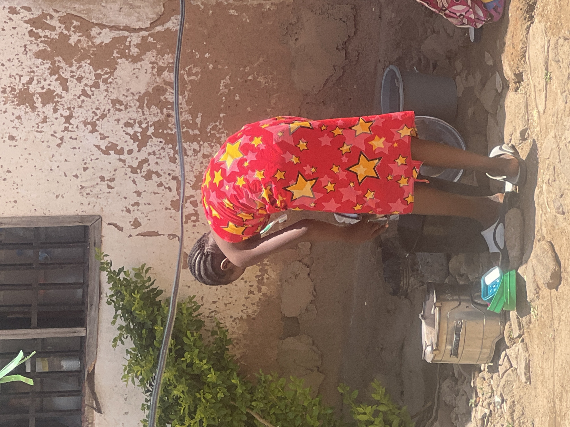 This image captures an outdoor scene in Gura Top, Nigeria, depicting a young female engaged in a domestic chore under bright daylight.
The main subject is a young female with dark skin and braided hair, dressed in a vibrant red garment adorned with numerous yellow and pink stars. She is bent forward at the waist, with her hands immersed in a container on the ground, suggesting she is washing something, possibly her hands, face, or small items. She wears light-colored sandals on her feet.
The setting appears to be a yard or courtyard. In the background, a wall with peeling paint or plaster reveals a darker, reddish-brown surface beneath. To the left, a window with a dark metal security grate is visible, partially obscured by green leafy plants or bushes that also grow along the ground. On the ground around the person, there are several utilitarian objects: two grey plastic buckets, one clear bucket, a metallic pot or container, and a small blue and green container. The ground itself is uneven, composed of dirt and scattered rocks.
The strong, direct sunlight indicates it is daytime, casting clear shadows and illuminating the scene brightly. There is no visible text in the image.