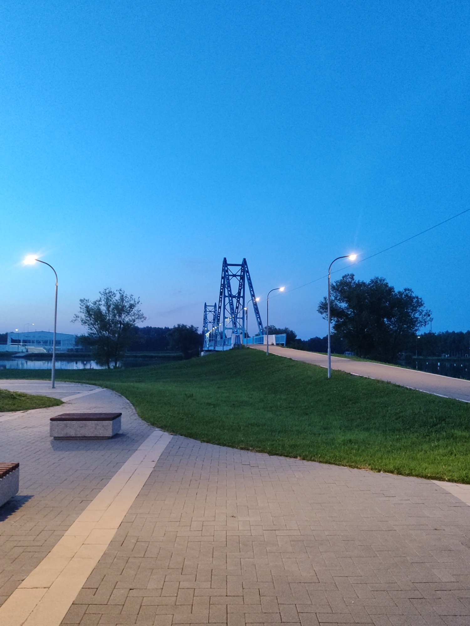 The image depicts a park-like setting at dusk in Bronnitsy, Russia. A pedestrian bridge with a distinctive blue metal truss structure spans across a body of water, possibly a river or lake, in the background. The bridge is illuminated by streetlights, as are the pathways leading up to it. The foreground features a paved walkway made of interlocking grey bricks with a contrasting lighter strip. Two stone benches with wooden seats are visible along the walkway. The area is landscaped with green grass. In the distance, across the water, there are trees and some buildings, one of which appears to be an industrial structure. The sky is a deep, clear blue, indicating the transition from daylight to night. There are no people visible in the scene. The overall atmosphere is serene and calm.